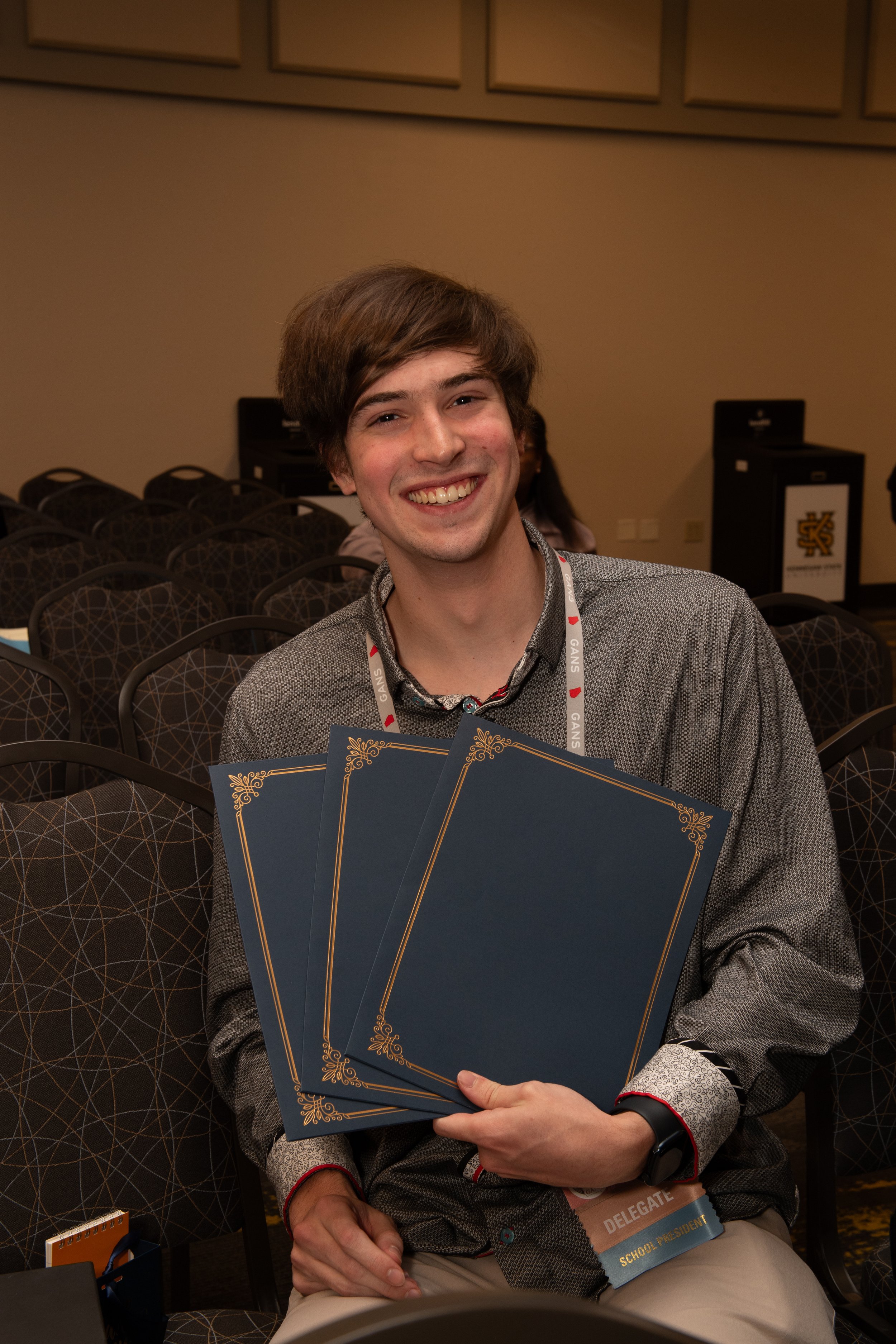 A young man with brown hair smiling and holding several certificates at a conference. He wears a gray shirt and has a delegate badge that says 'School President.' The conference room has chairs and a podium in the background.