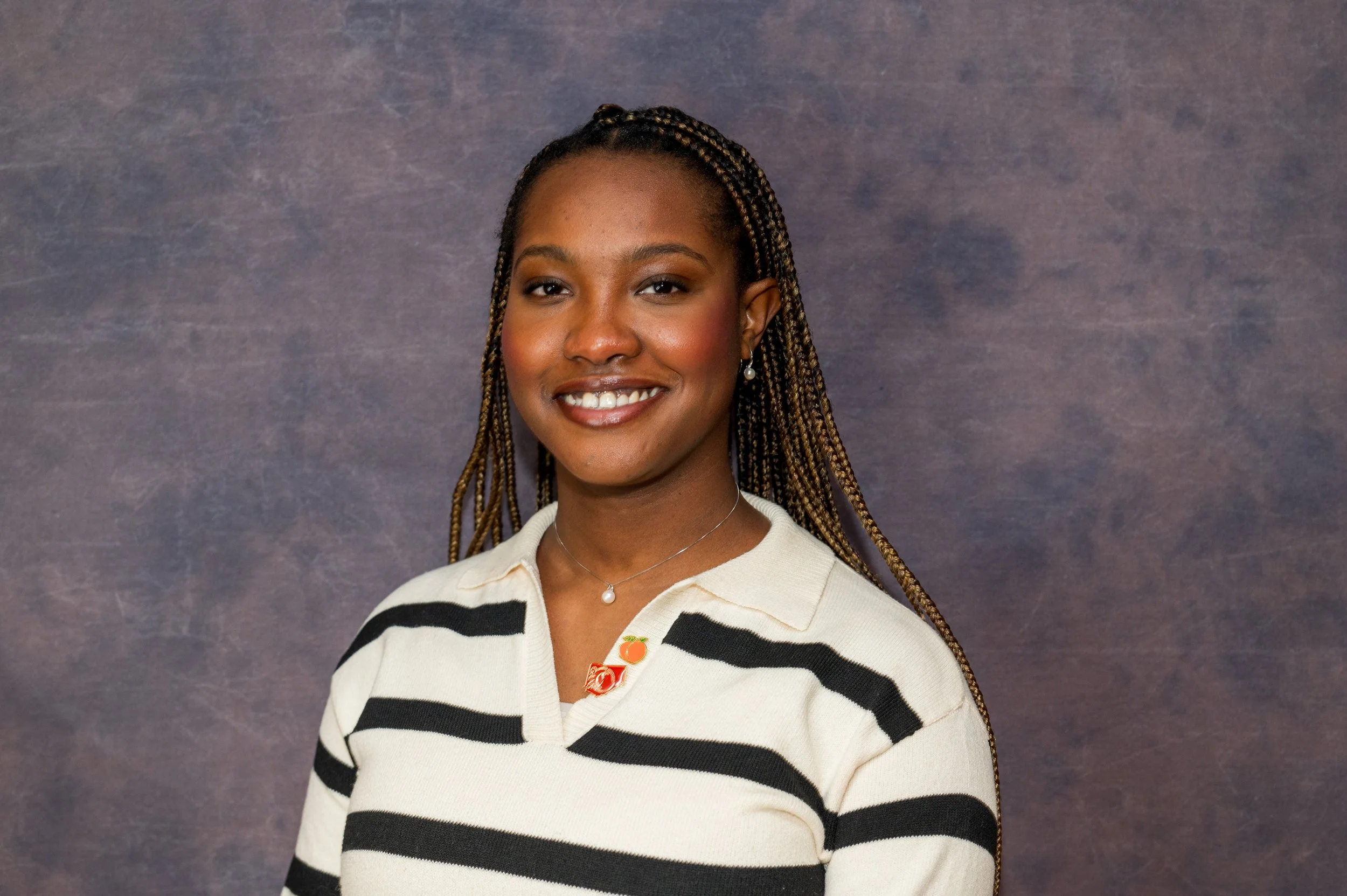 A young woman with braided hair and earrings standing against a gray background, smiling at the camera.