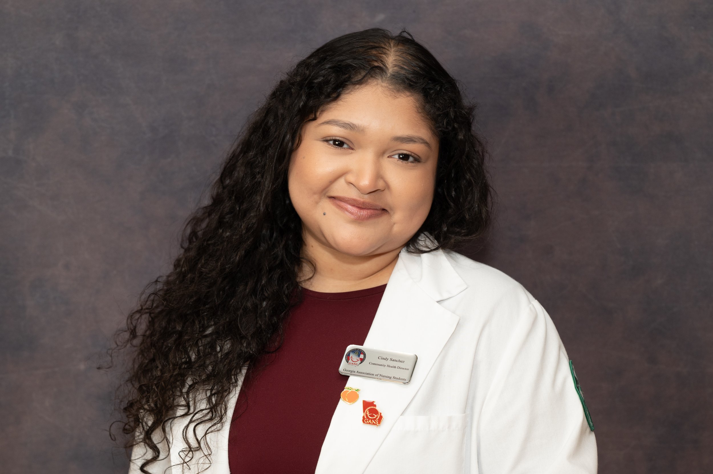 Portrait of a woman with long curly hair wearing a white lab coat and a name badge, standing against a dark background.