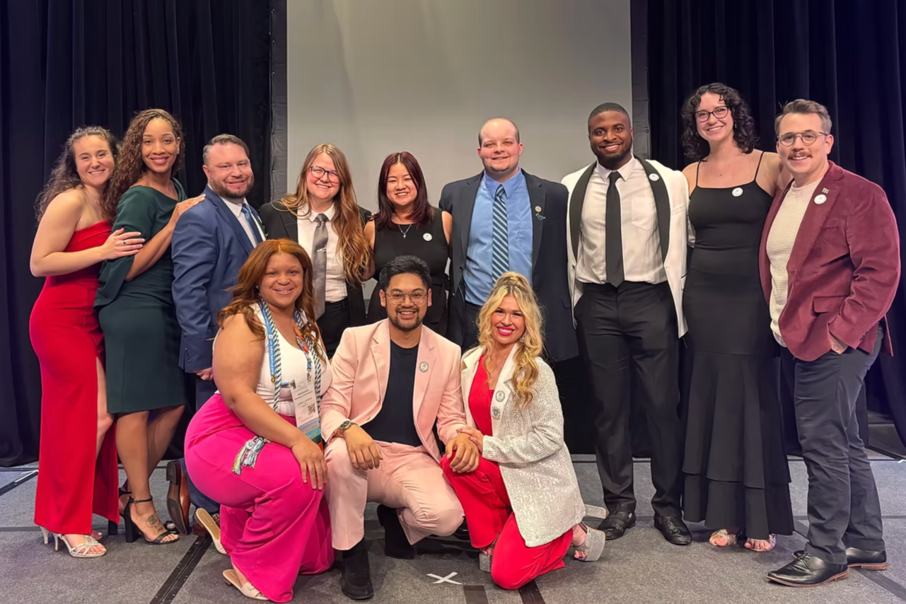 Group of people dressed in formal and semi-formal attire, posing together on stage at an event or conference.
