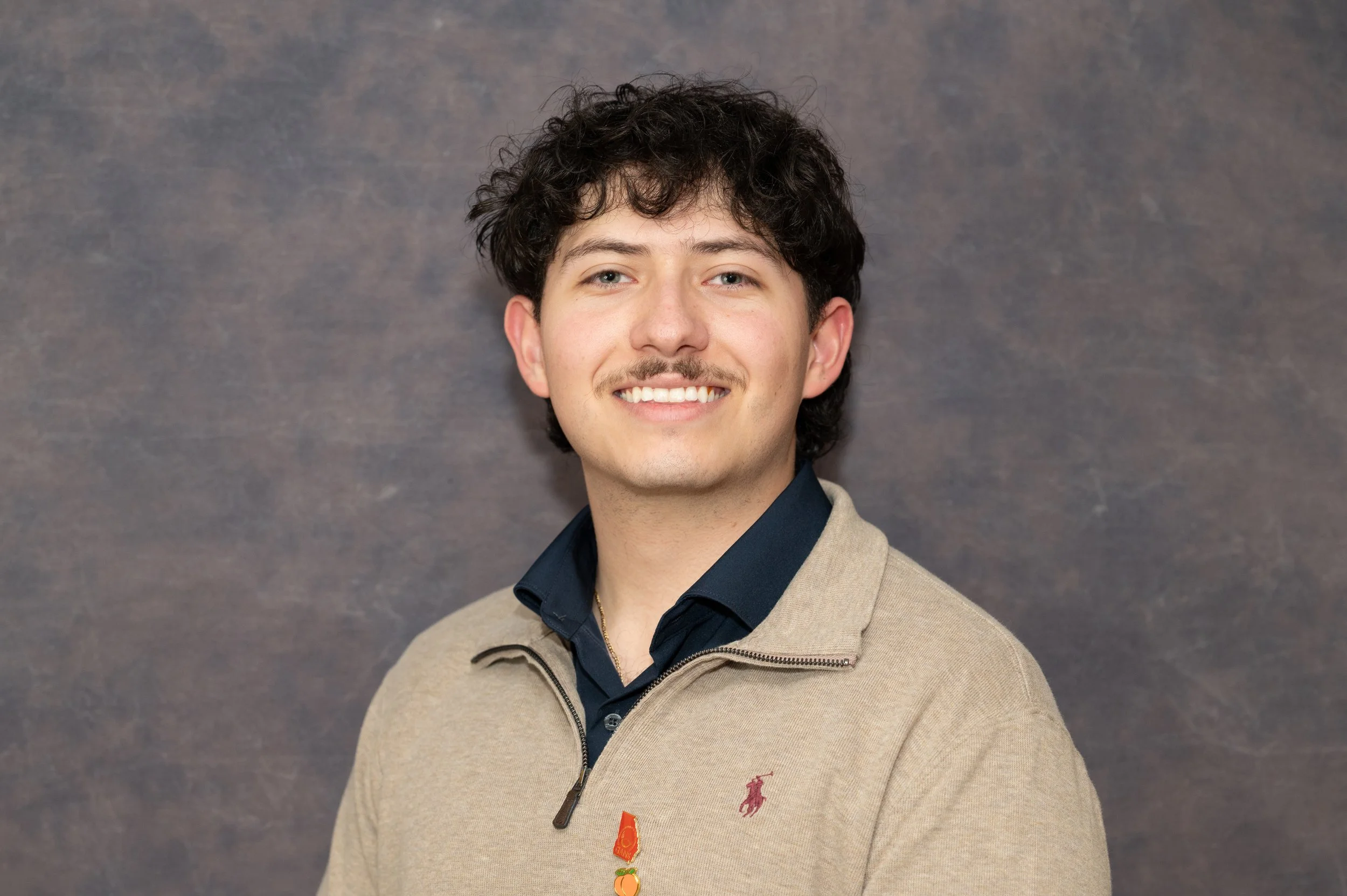 A young man with dark curly hair and light skin, smiling, wearing a beige zip-up jacket with a small red logo on the chest, a dark collared shirt underneath, and a necklace with cartoon characters against a gray mottled background.
