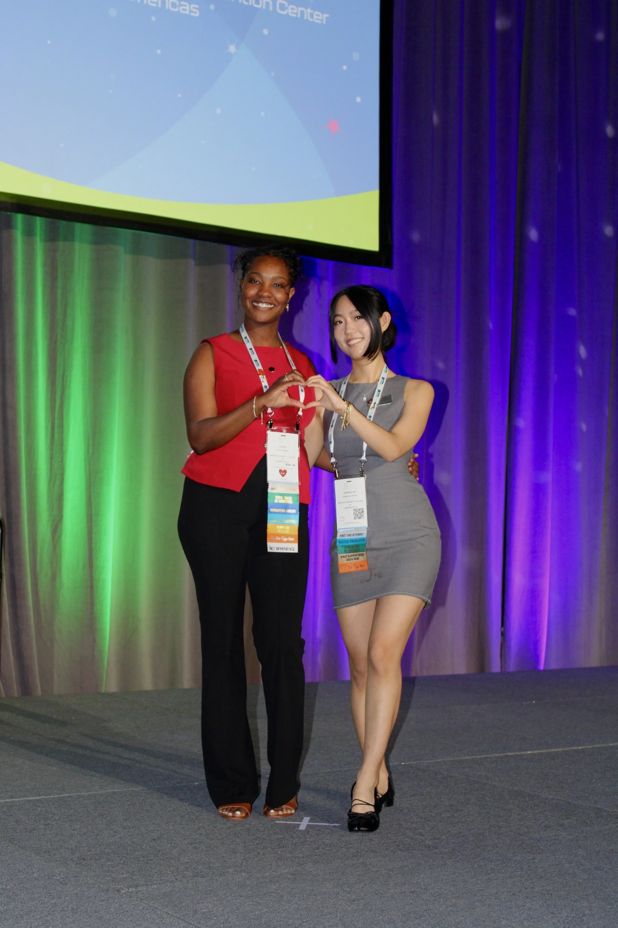 Two women standing on stage making a heart shape with their hands, smiling. Behind them is a large screen and colorful curtains, suggesting a conference or event.