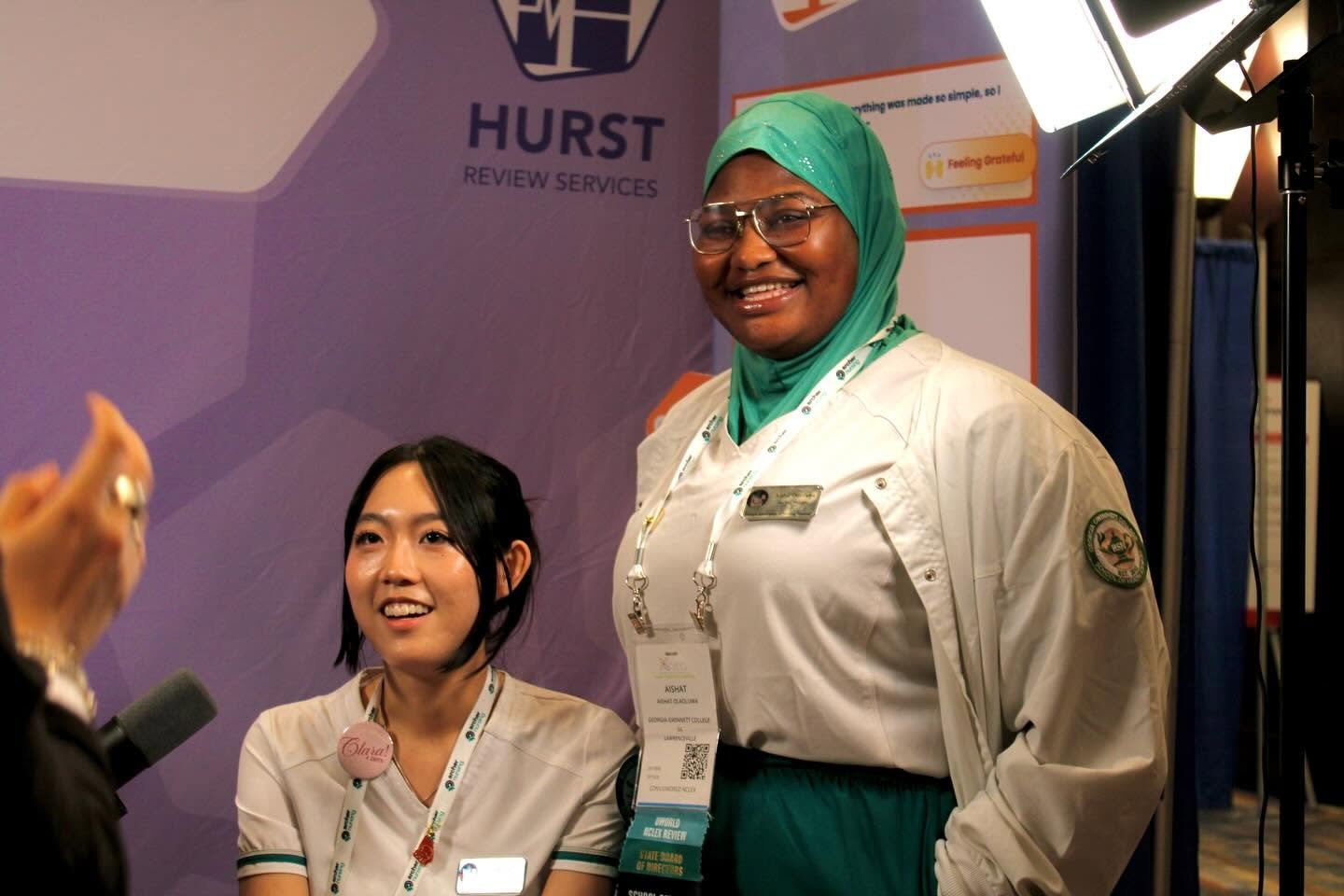 Two women, one sitting and one standing, at a booth with Hurst Review Services branding, engaging in a conversation with someone outside the frame in a professional setting.