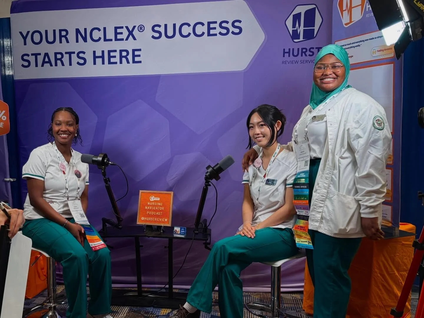 Four healthcare professionals in scrubs and white coats smiling at a medical conference or event, seated and standing in front of a purple backdrop with the words "Your NCLEX Success Starts Here" and logos, with microphones, badges, and a sign that reads "Nursing Educator Podcast."