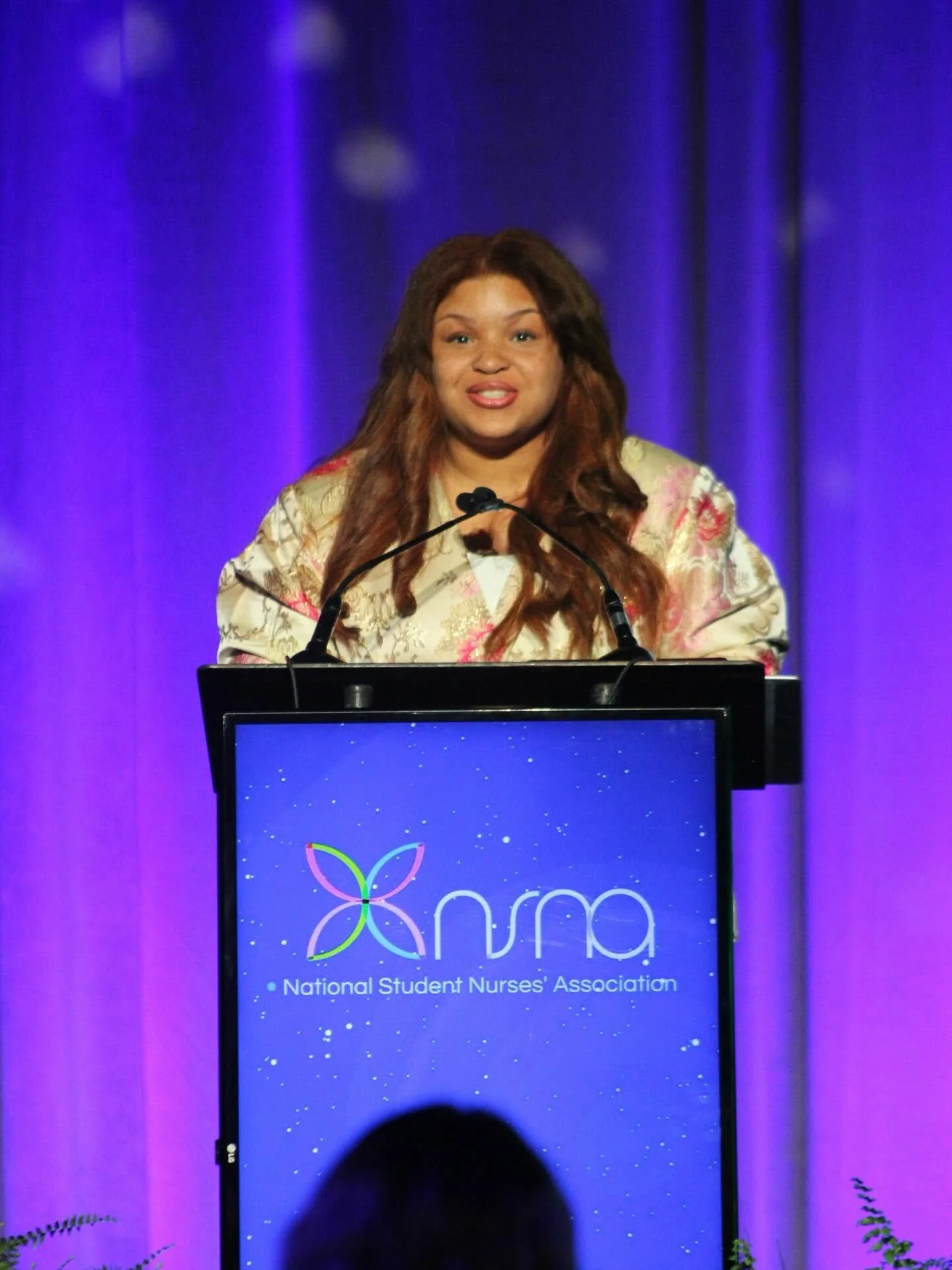 A woman is speaking at a podium with the logo of the National Student Nurses' Association on it, against a purple curtain backdrop.