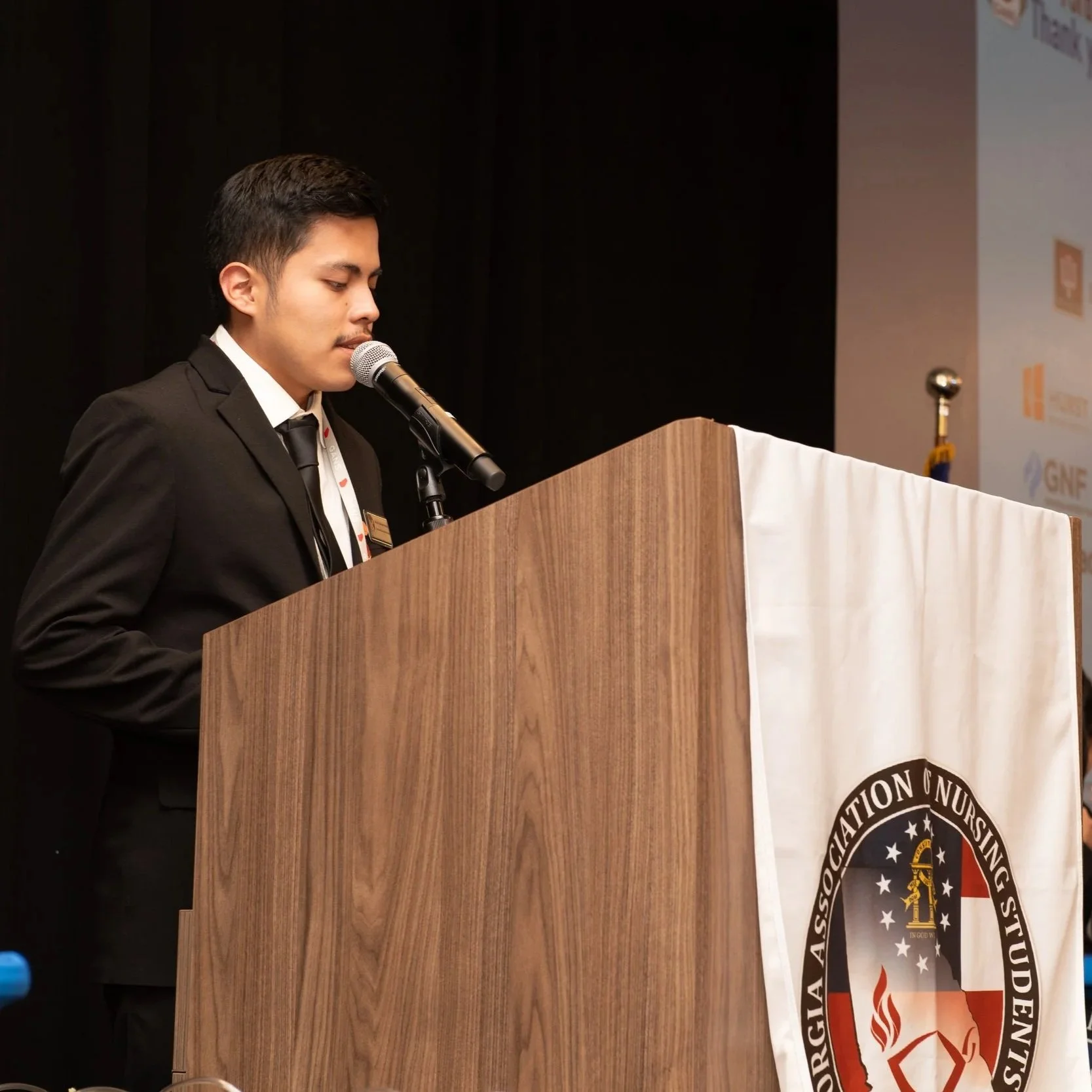 Young man in formal attire standing at a wooden podium with a microphone delivering a speech at an event, with a banner displaying the logo of an organization behind him.
