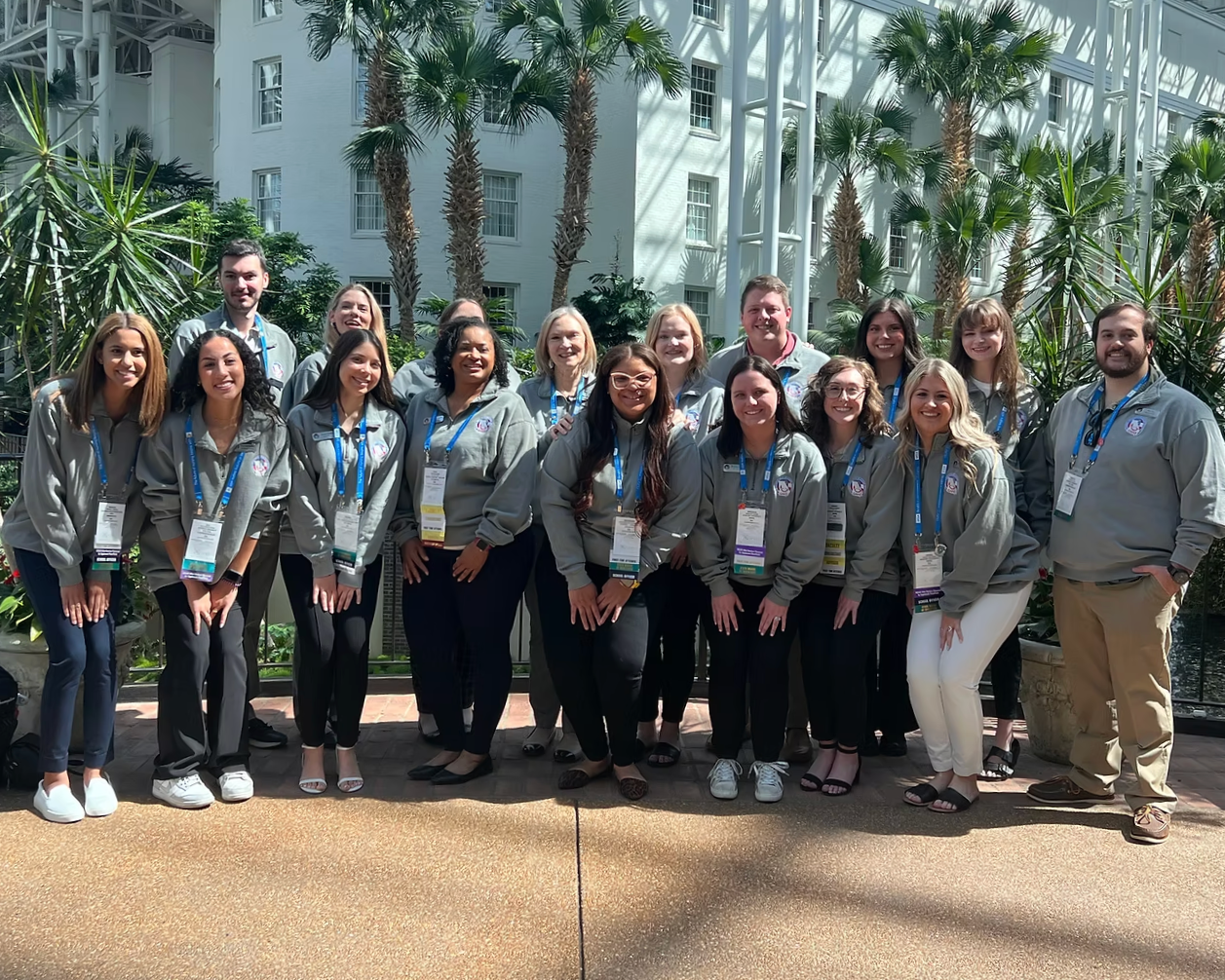A group of young adults posing outdoors in front of tropical plants and a white building, all wearing gray jackets and conference badges.