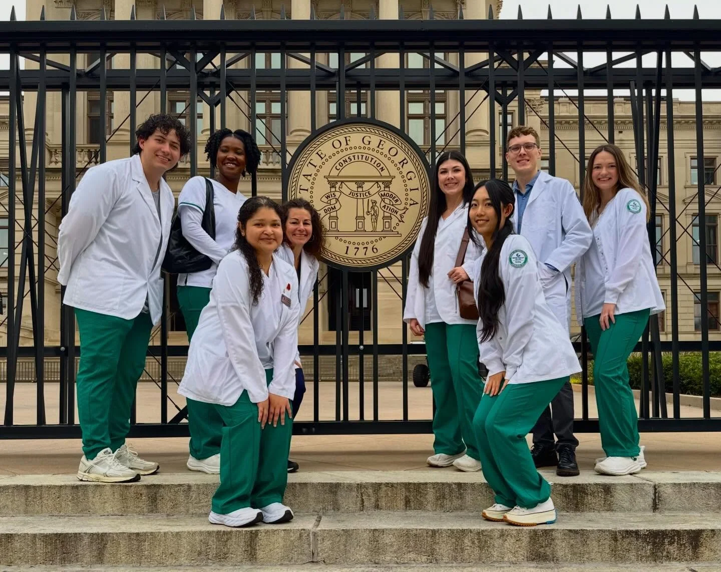 Group of nine students wearing medical scrubs and white coats, standing on steps in front of the Georgia state seal behind a gate outside a government building.