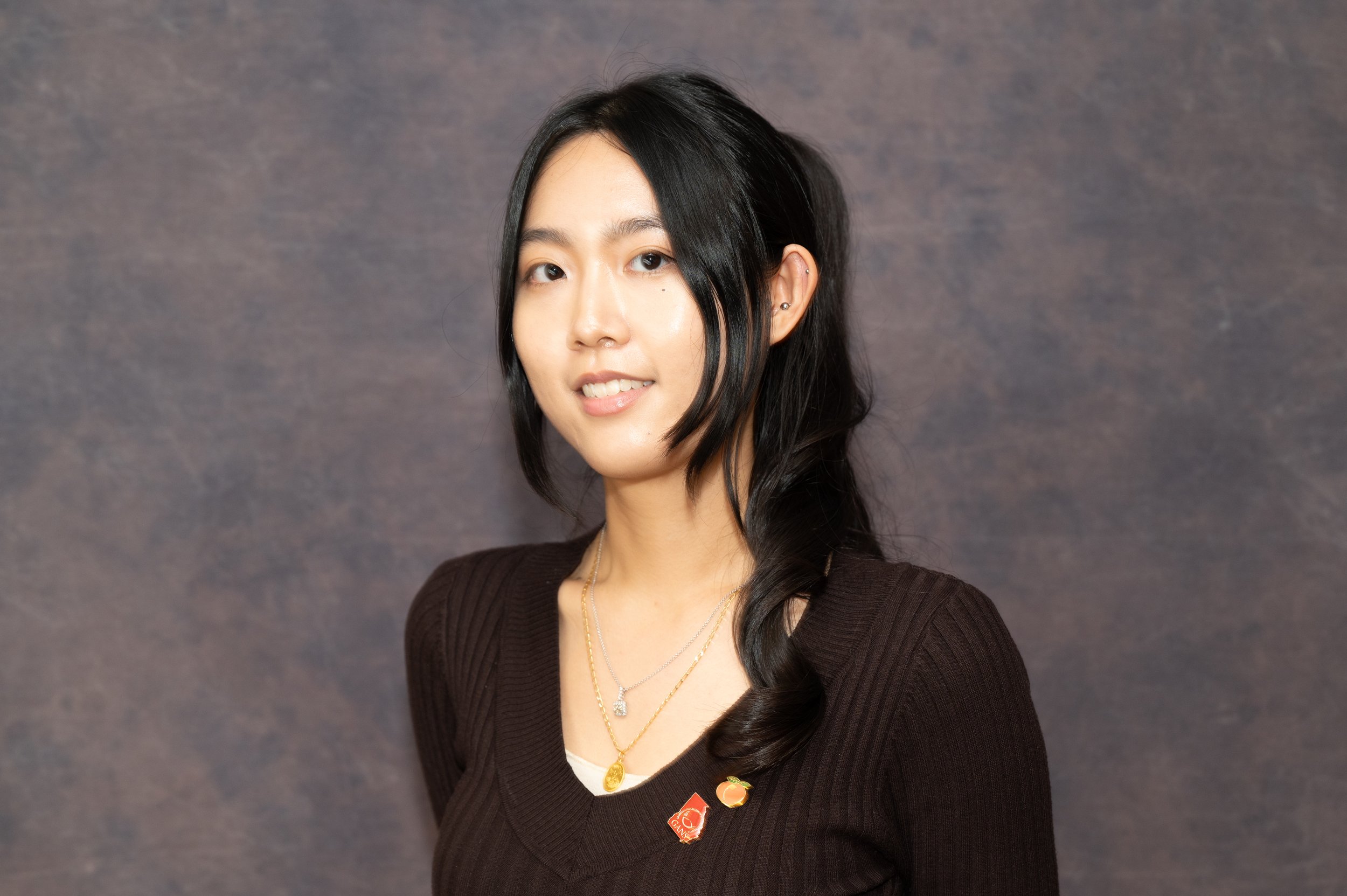 A woman with long black hair and multiple earrings, wearing layered necklaces and a brown top with pins, smiles slightly against a dark gray background.