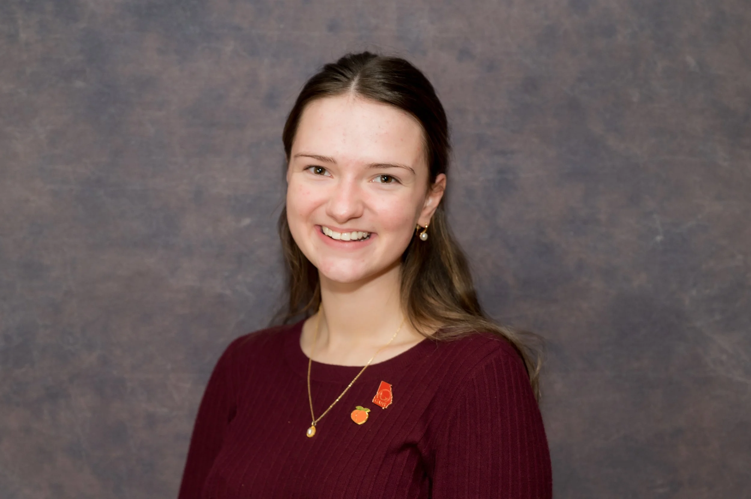 A young woman with brown hair and a warm smile, wearing gold earrings and a maroon sweater, standing against a textured gray background.
