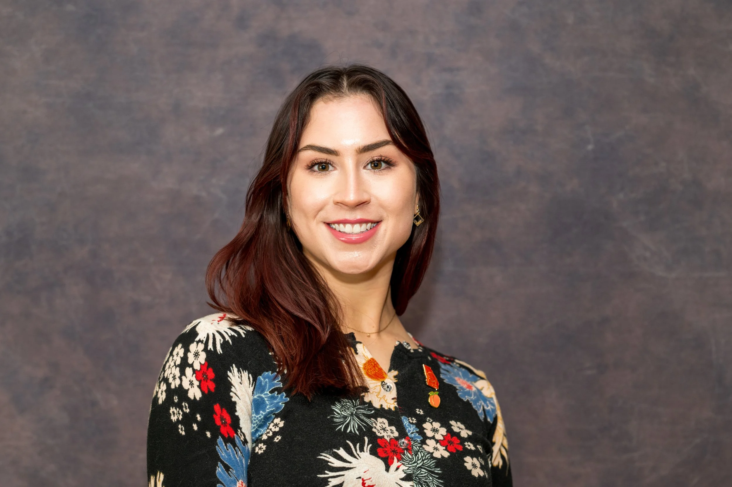 A woman with long brown hair, wearing a colorful floral patterned top, smiling, against a gray textured background.