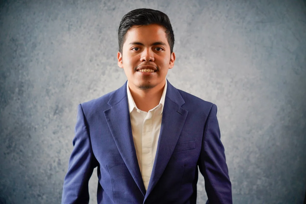 A young man with dark hair, wearing a blue suit and white shirt, smiling while standing against a gray textured background.