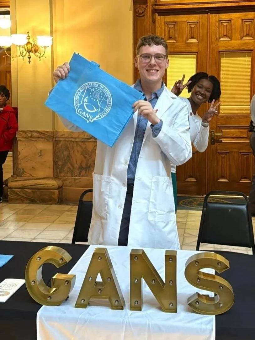 A young man in a white coat holding a blue flag with a logo. Behind him, a woman makes a peace sign. There is a table in front with gold letter decorations spelling 'GANS'. The setting appears to be an indoor event with wood-paneled walls and warm lighting.