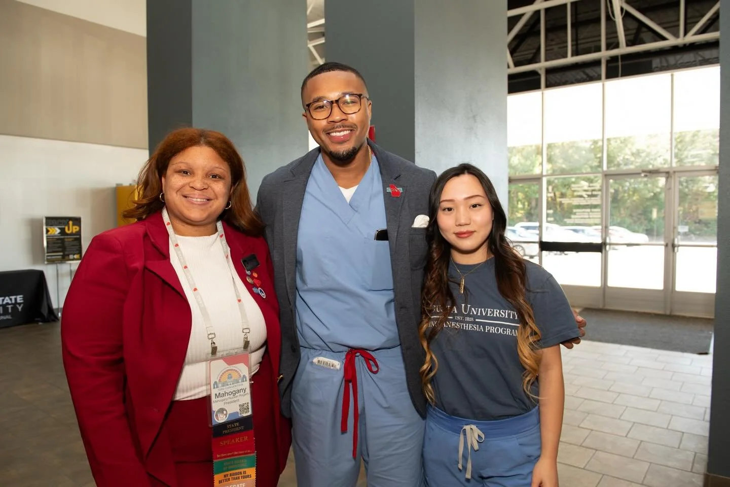 Three individuals standing together indoors, smiling at the camera. The woman on the left is wearing a red blazer and white top, with a name badge. The man in the middle is dressed in scrubs with a blazer over them. The young woman on the right is wearing a navy T-shirt and blue shorts, with a name badge that indicates she is part of a university program.