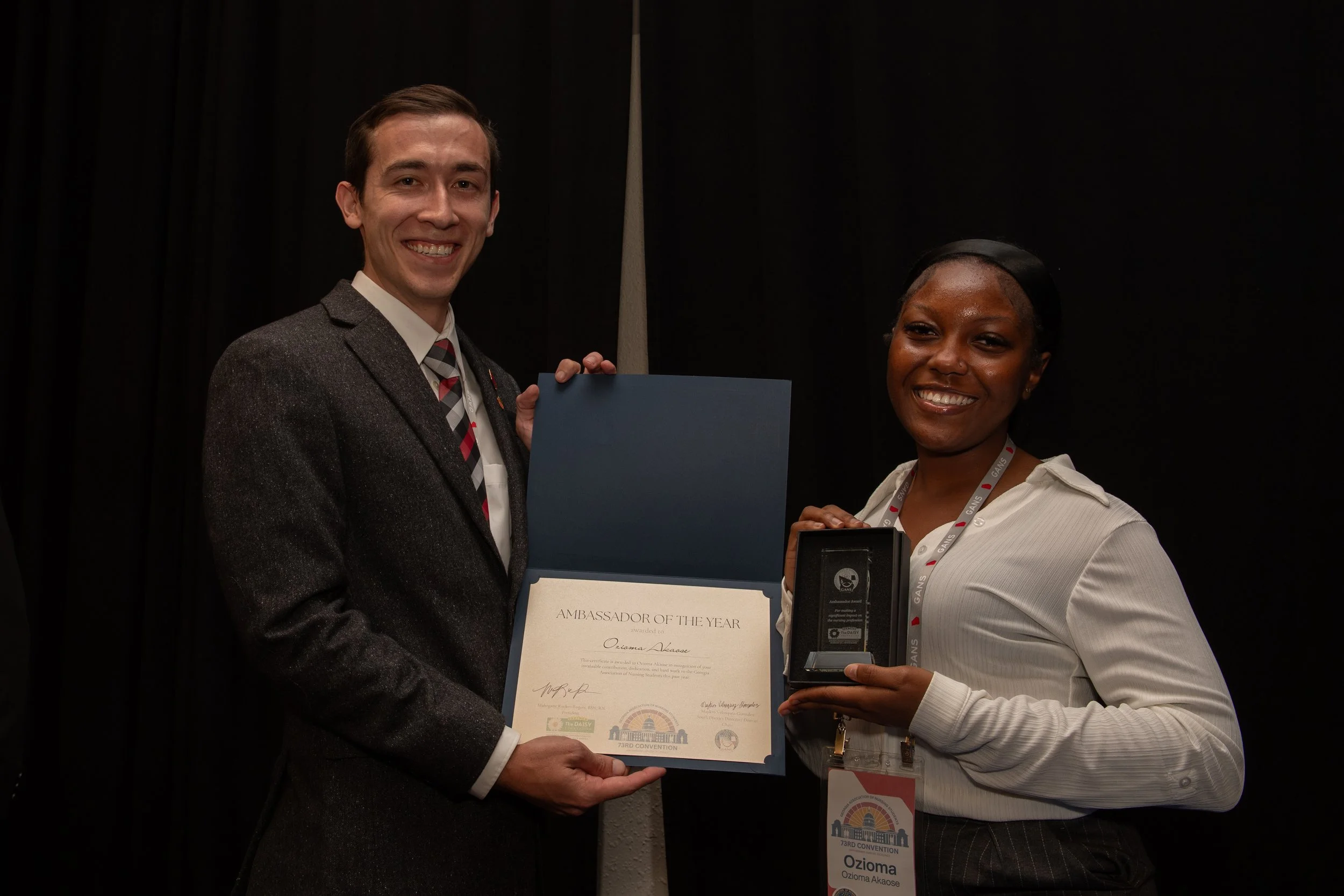 Two people standing together, smiling, with one holding a certificate that says "Ambassador of the Year" and the other holding a box with an award inside. They are at a formal event with black curtains in the background.