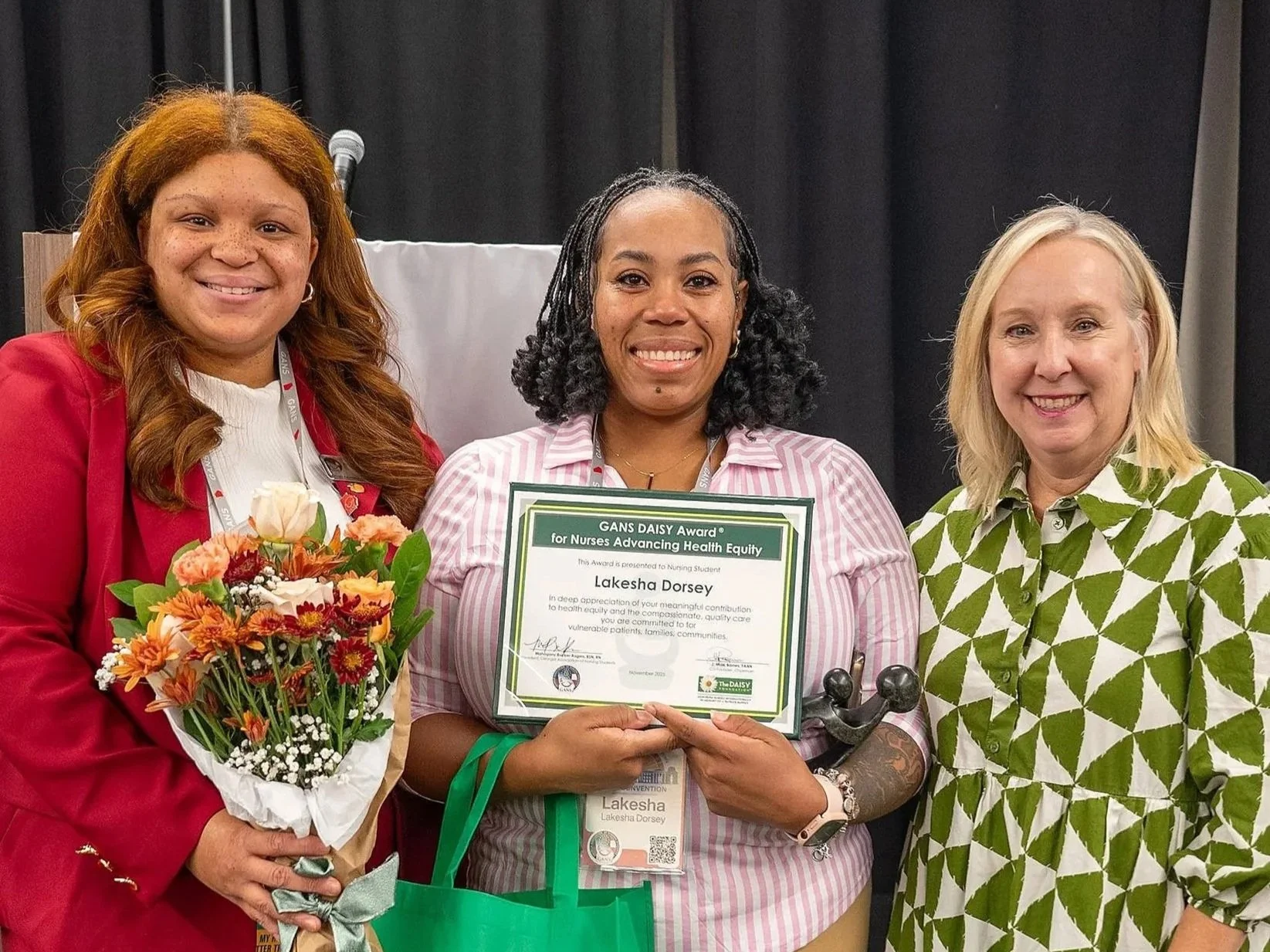 Three women standing together, center woman holding a certificate, background with dark curtains, the woman on the left holding a bouquet of flowers, and the woman on the right wearing a patterned green and white shirt.