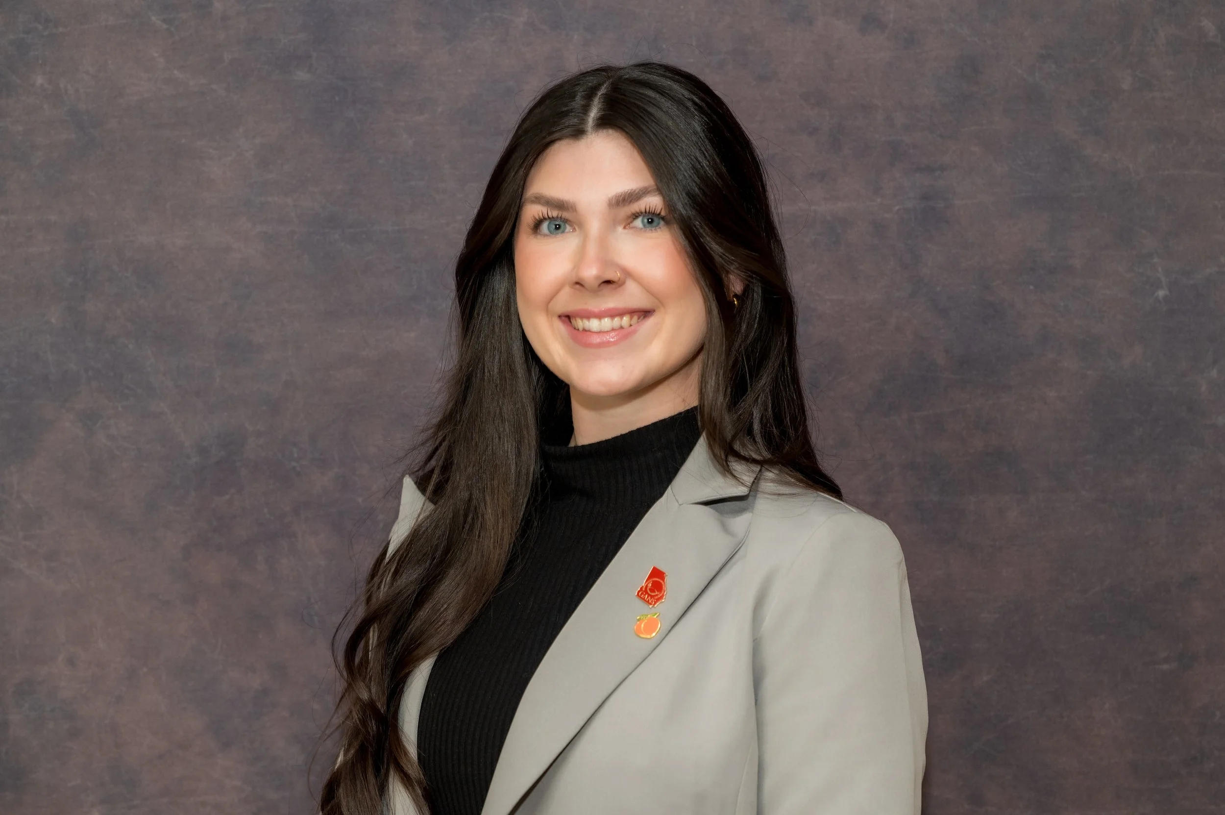 A young woman with long dark hair, blue eyes, and a nose ring smiling at the camera, wearing a black turtleneck and a light gray blazer, against a textured dark background.
