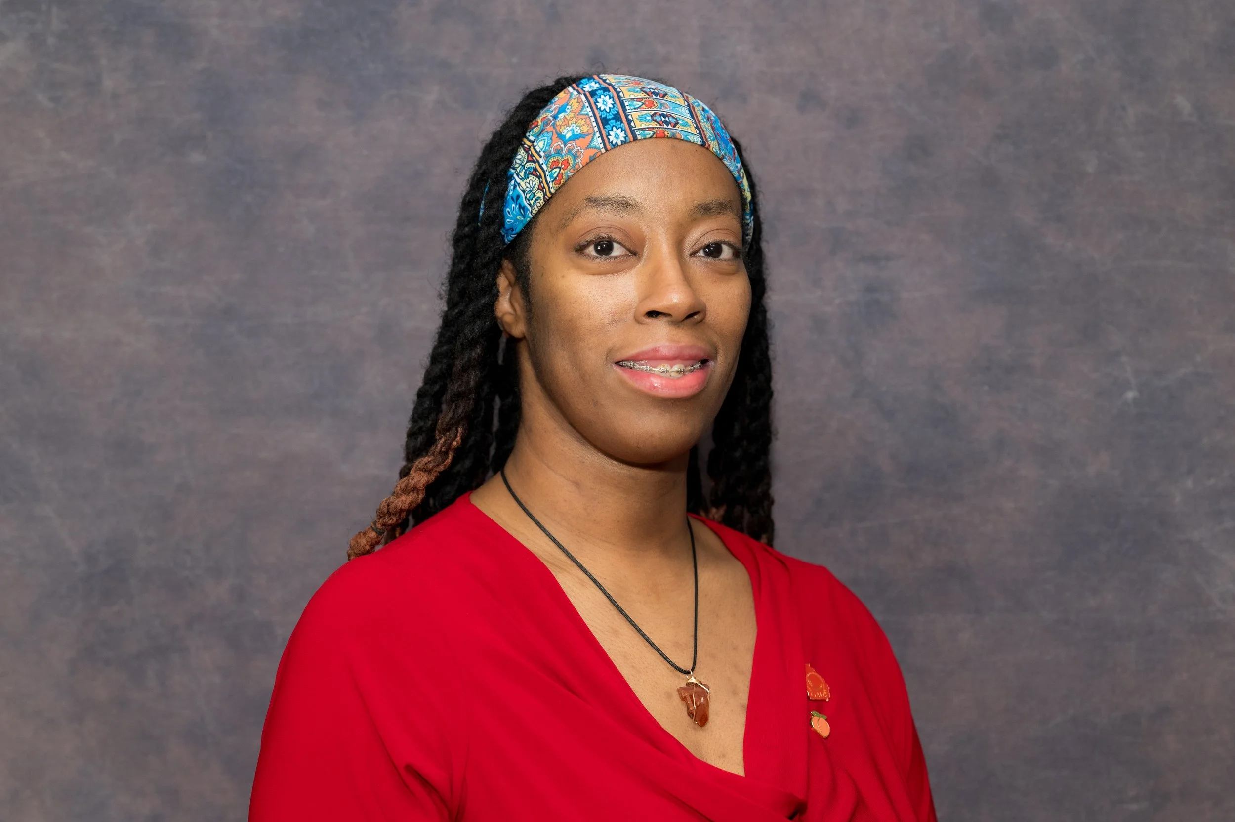 A woman with dark skin, wearing a colorful headscarf, a red top, and jewelry, standing against a gray background.