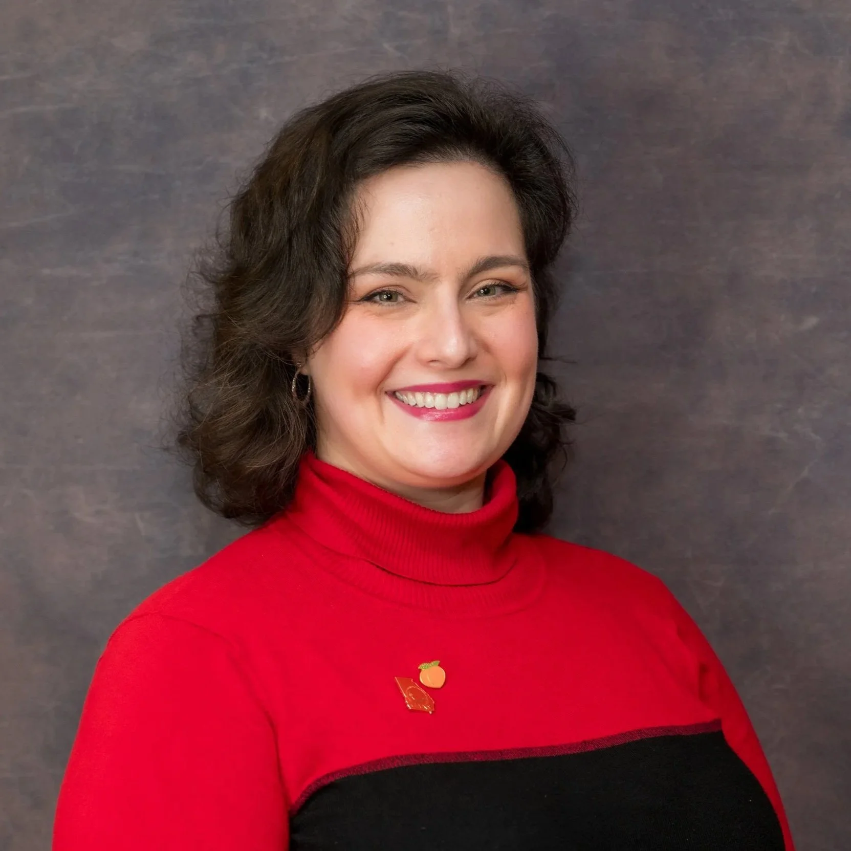 A woman with dark brown, curly hair smiling and wearing a red turtleneck sweater, standing against a dark gray background.
