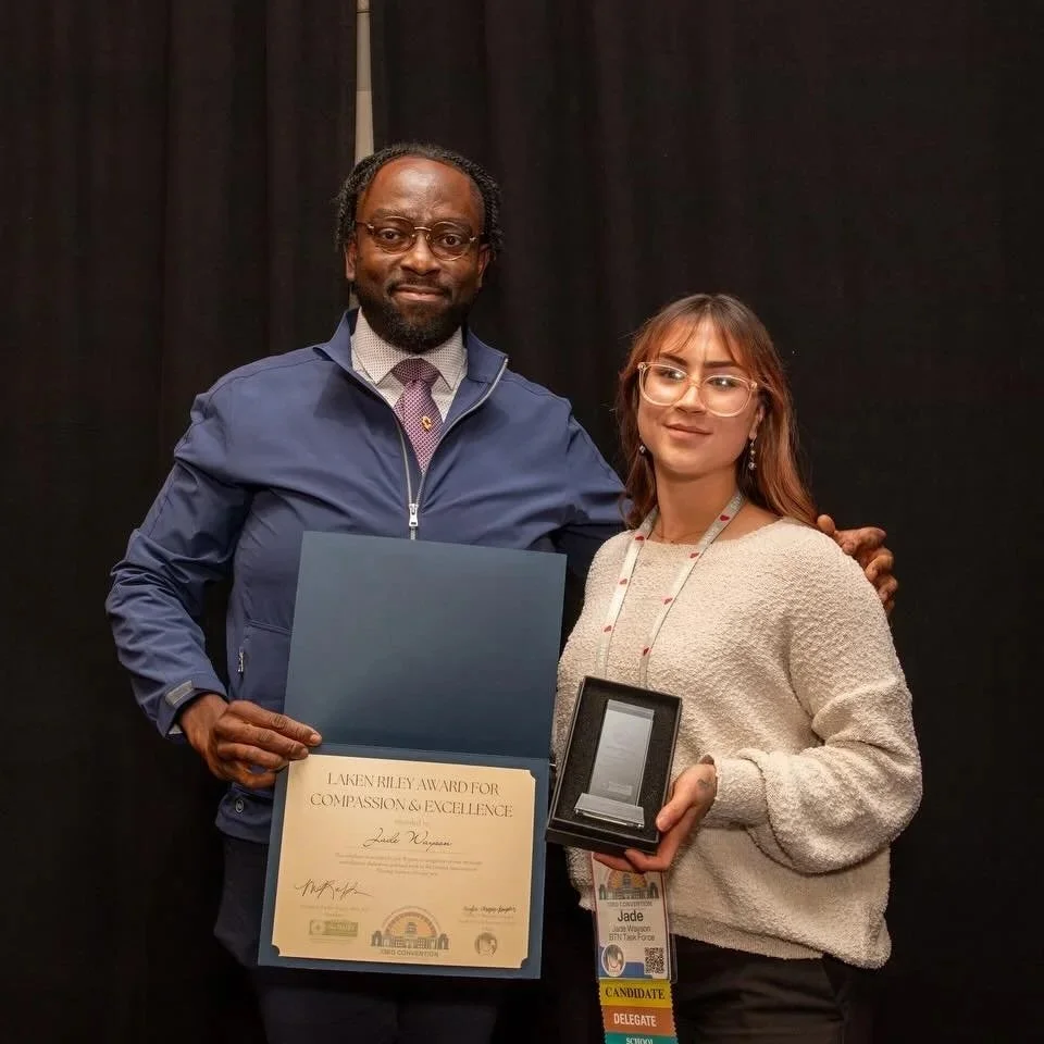 A man and a woman are standing together on stage. The man is holding a certificate that says 'Laken Riley Award for Compassion & Excellence,' and the woman is holding a box with an award inside. Both are smiling and dressed casually. The background is black.