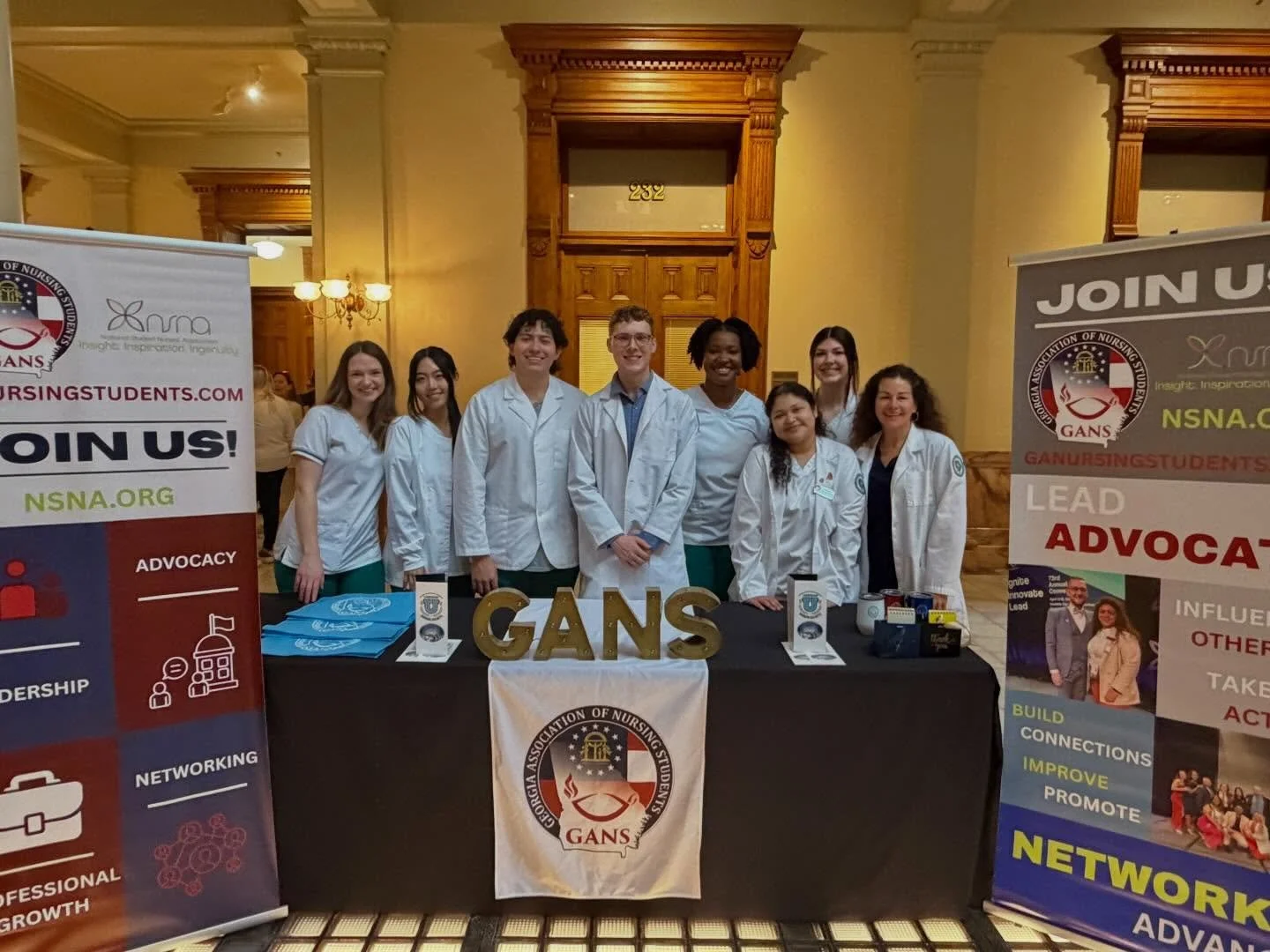 Group of nine diverse nursing students in white coats standing behind a table with educational materials at a professional event, with promotional banners on either side promoting nursing advocacy, leadership, and networking.