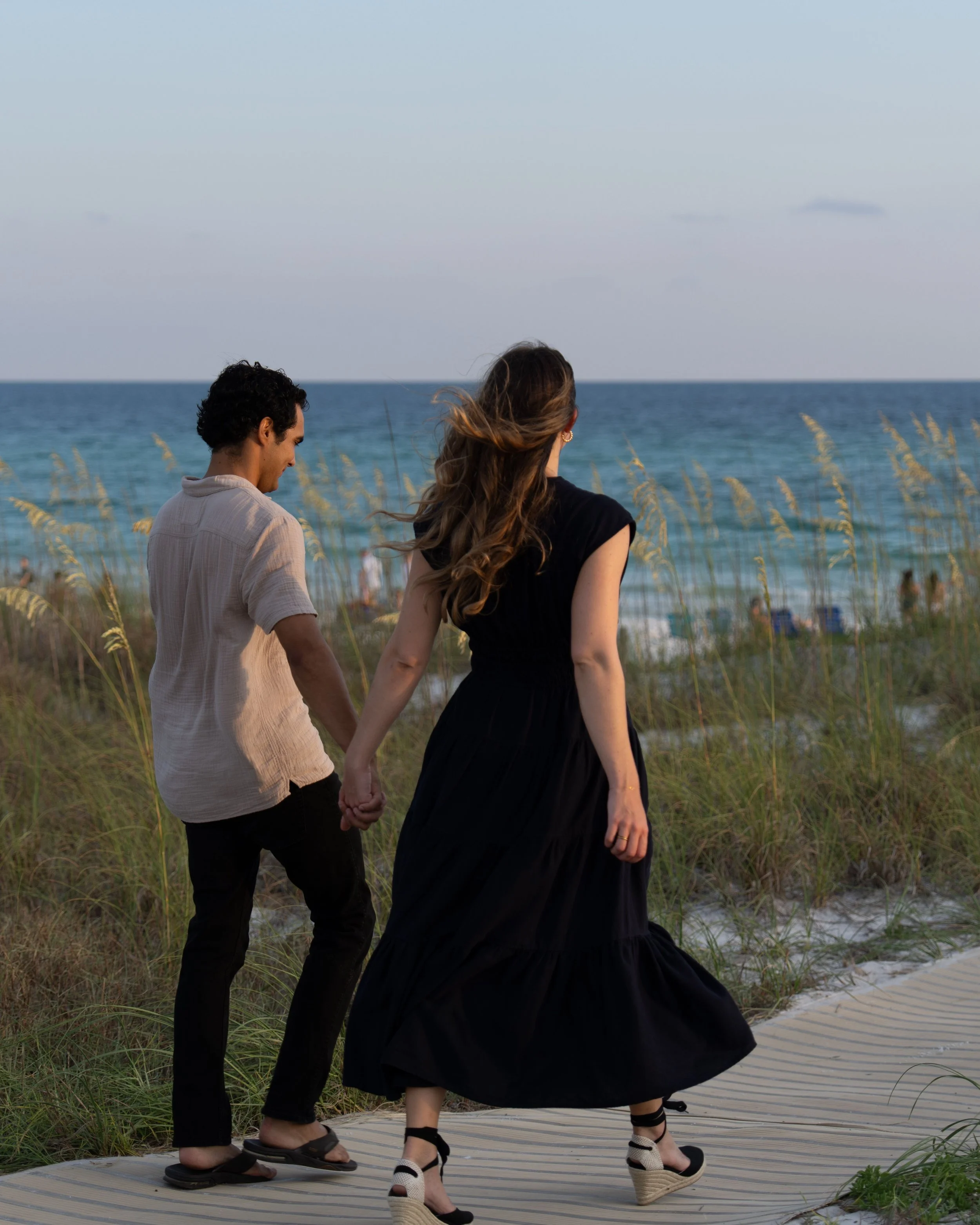 A couple holding hands walking on a wooden pathway at the beach, with the ocean and sky in the background.