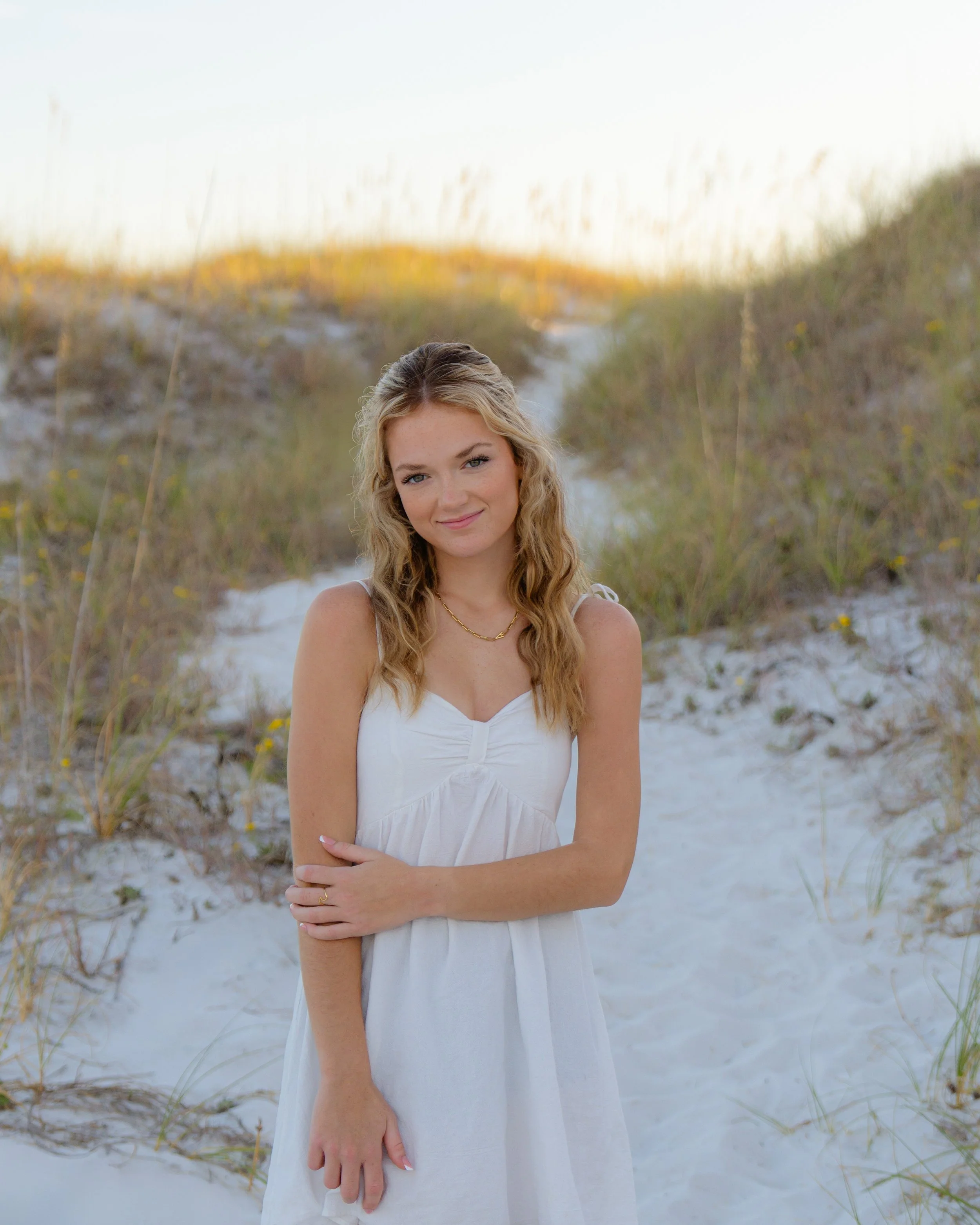 Young woman with long blonde hair standing on a sandy beach with grass-covered dunes in the background, wearing a white summer dress and gold jewelry.