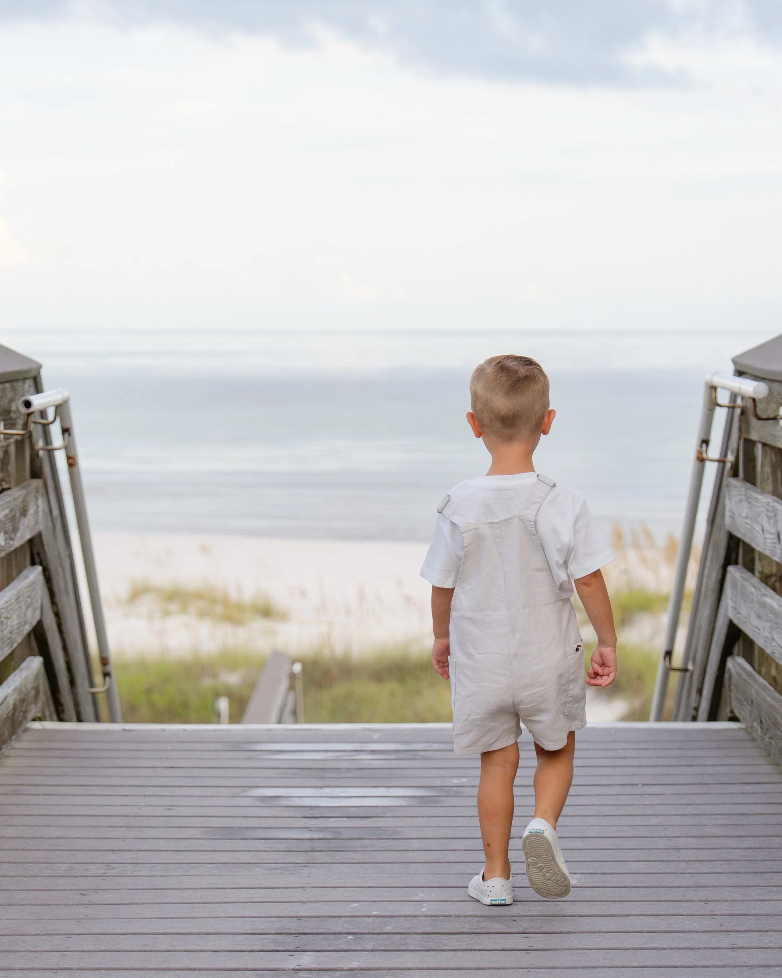 A young boy walking down a wooden ramp towards the beach, with the ocean and sky in the background.