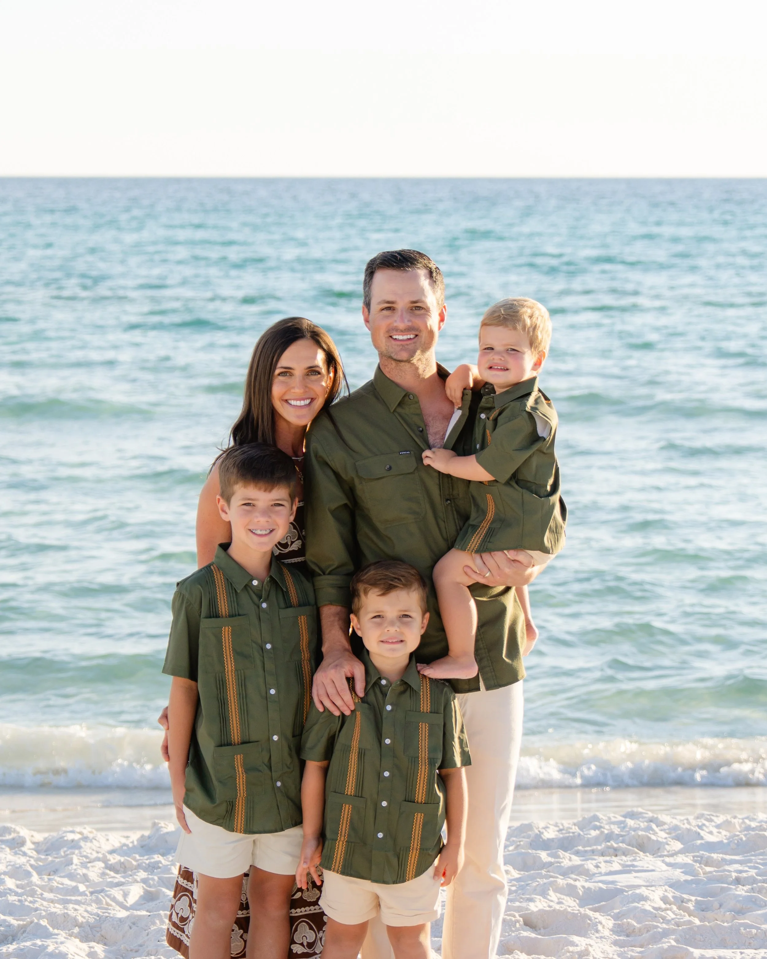 A family of five standing on the beach near the ocean, dressed in matching green shirts and white shorts, smiling at the camera.