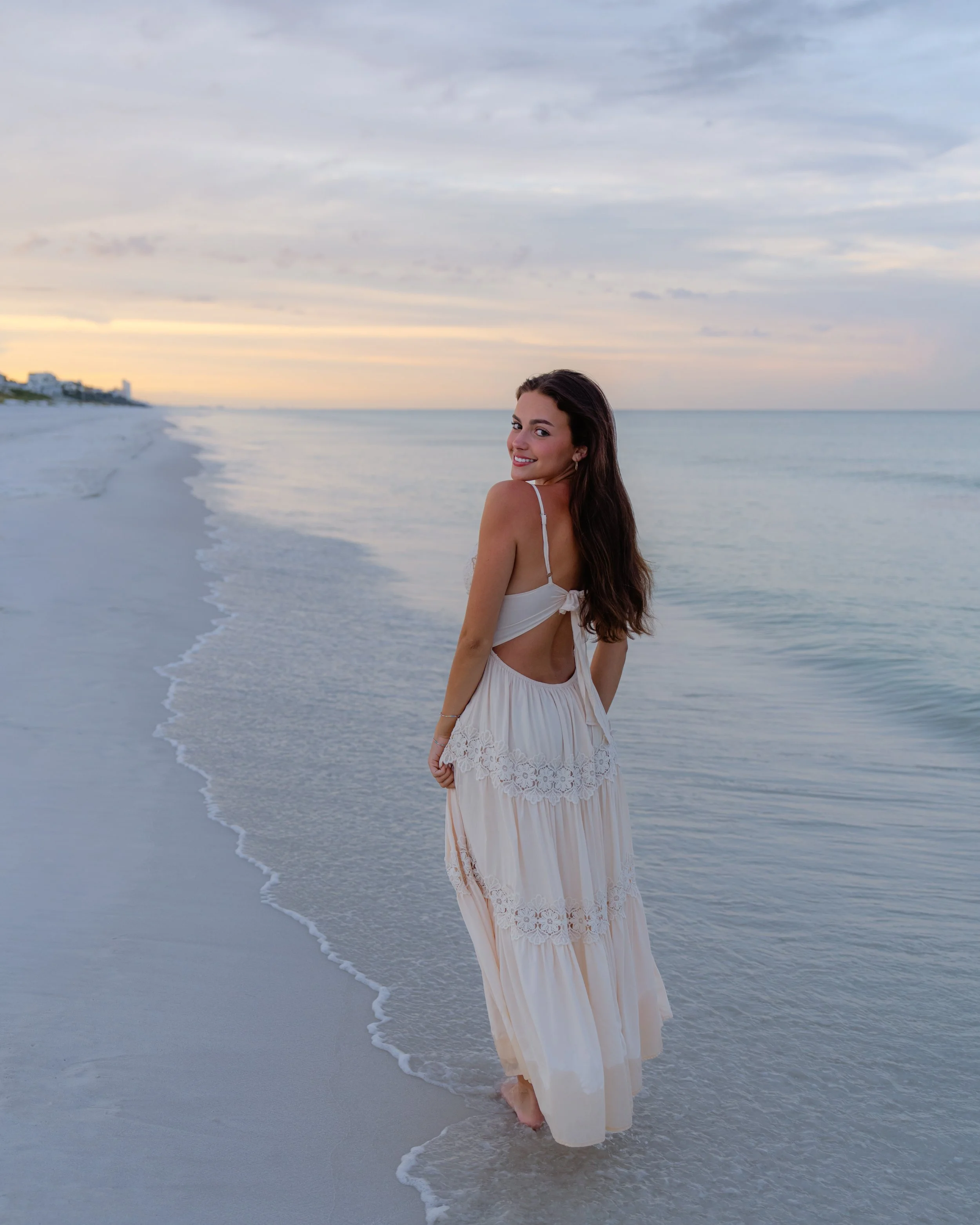 Young woman in a white dress walking on the beach at sunset, smiling at the camera.