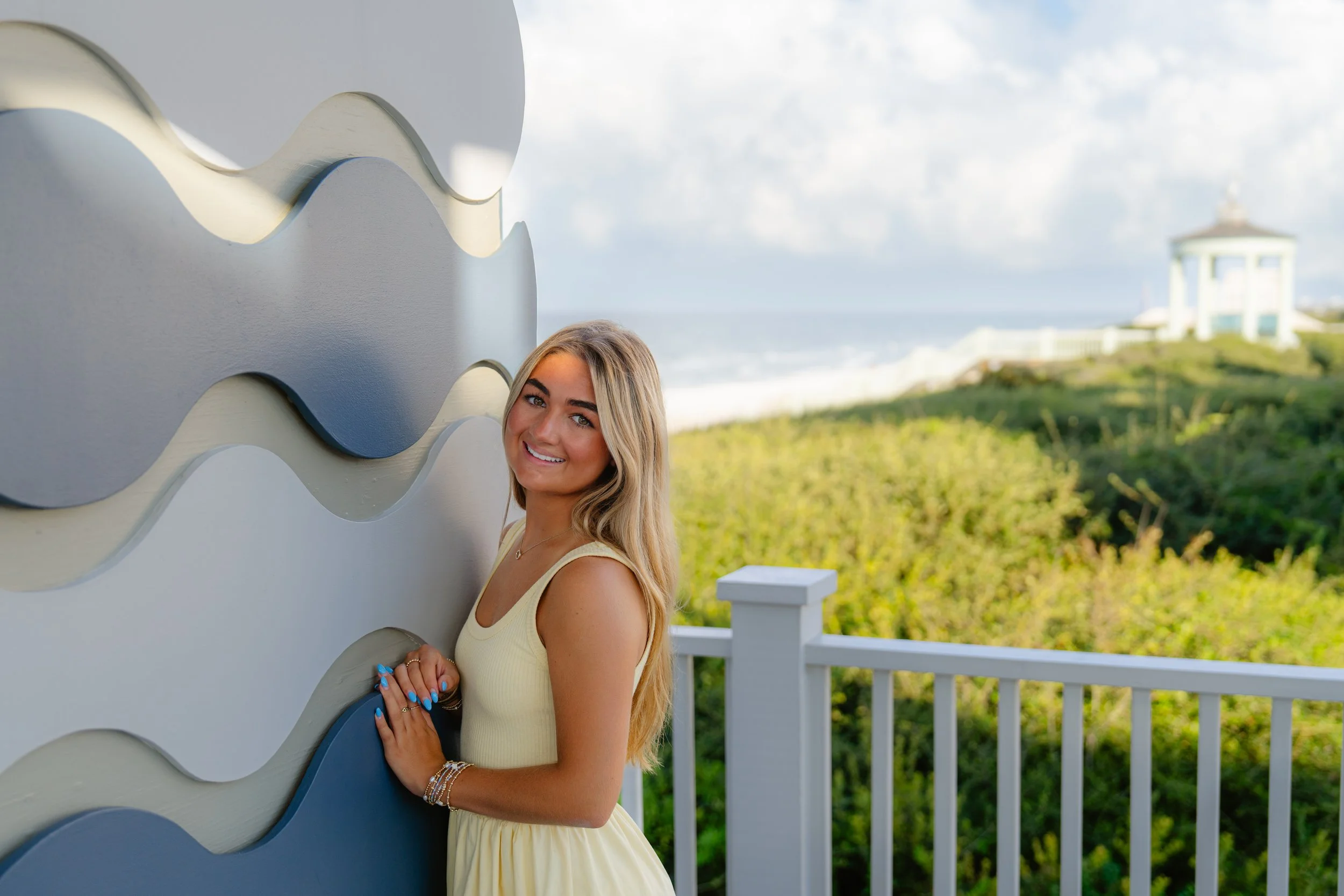 A young woman with long blonde hair smiling and leaning against a colorful wave-shaped wall outdoors near the beach, with greenery and a gazebo in the background.