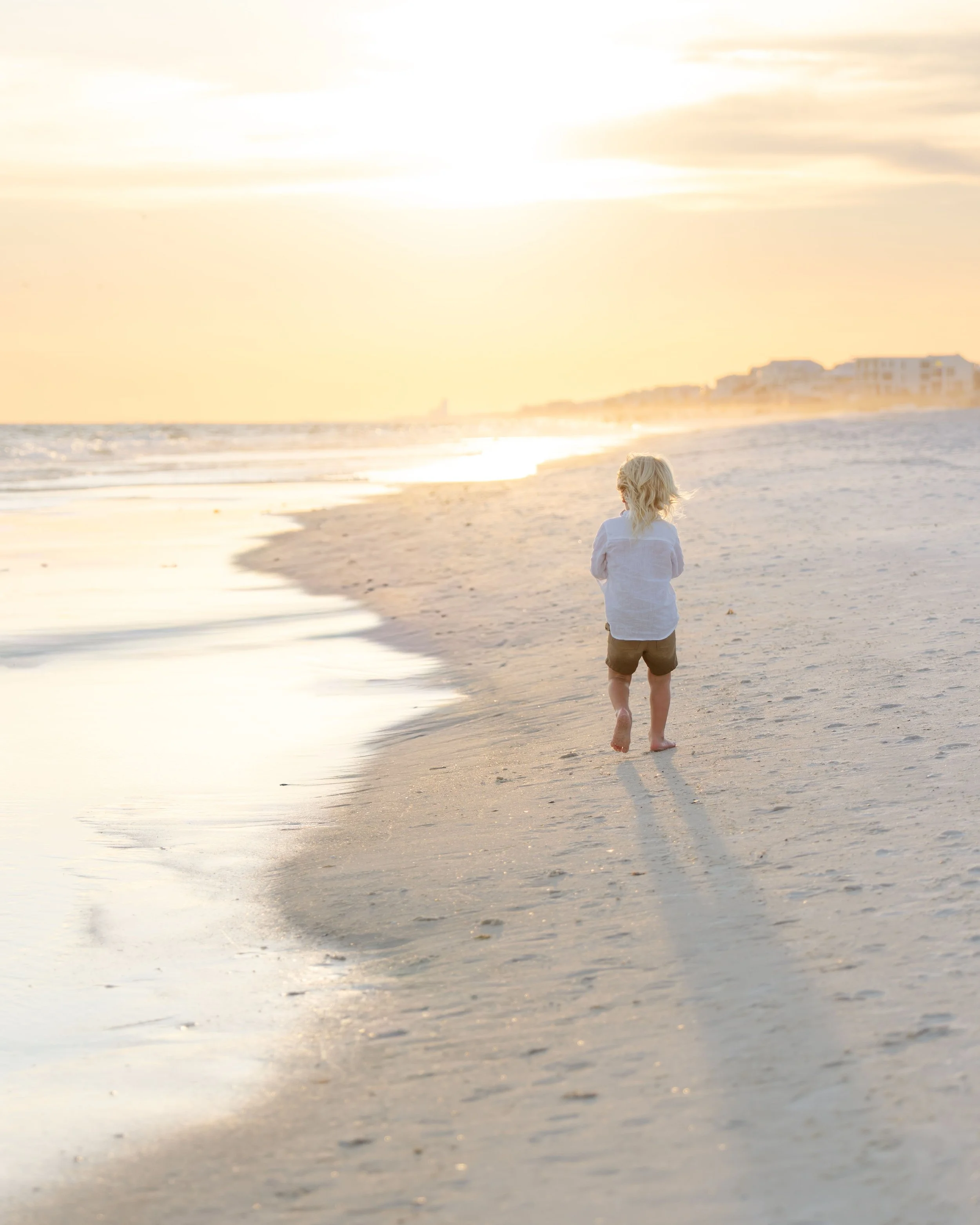 A young child with blonde hair wearing a white shirt and shorts walking alone on the sandy beach during sunset.