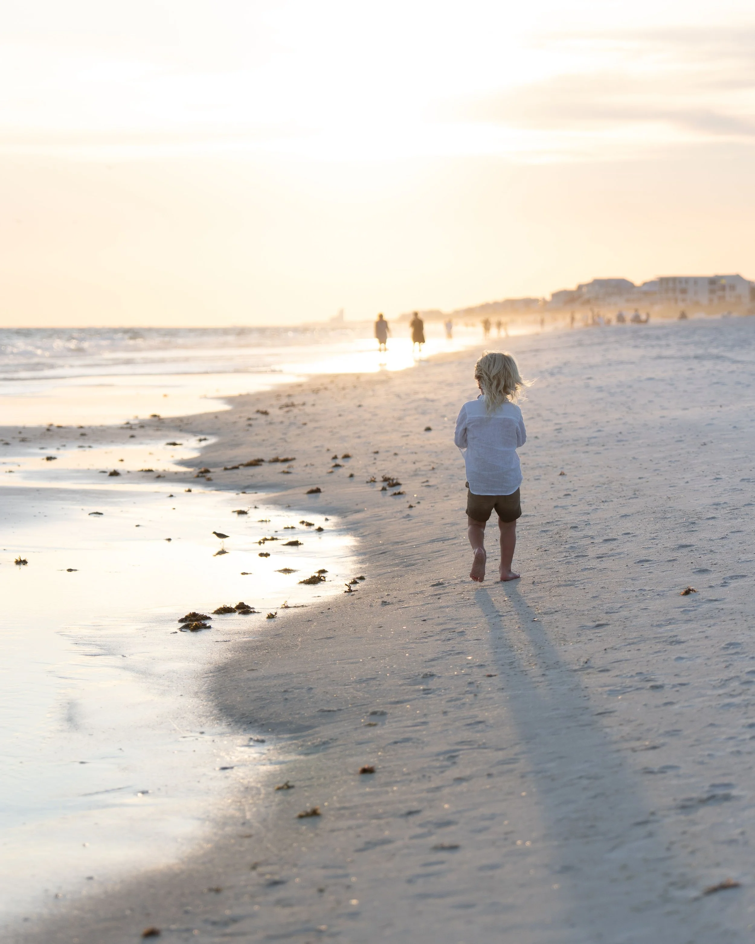 Child walking alone on sandy beach during sunset or sunrise, with other people in the distance and buildings along the shoreline.