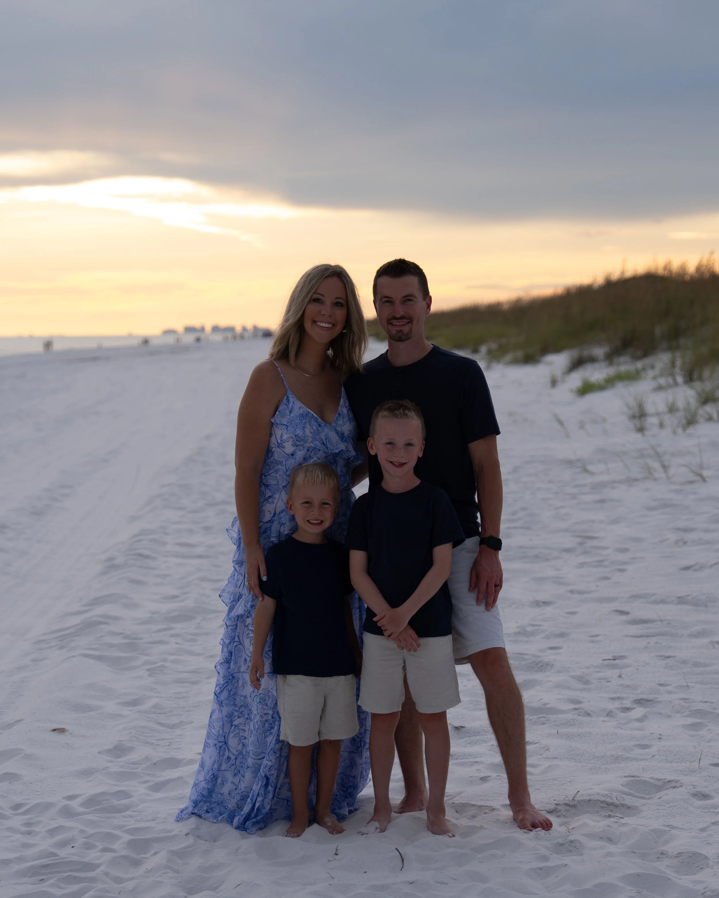 A family of four standing on the beach at sunset, with sand dunes and the ocean in the background. The mother, father, and two young sons are smiling and dressed in casual summer clothing.