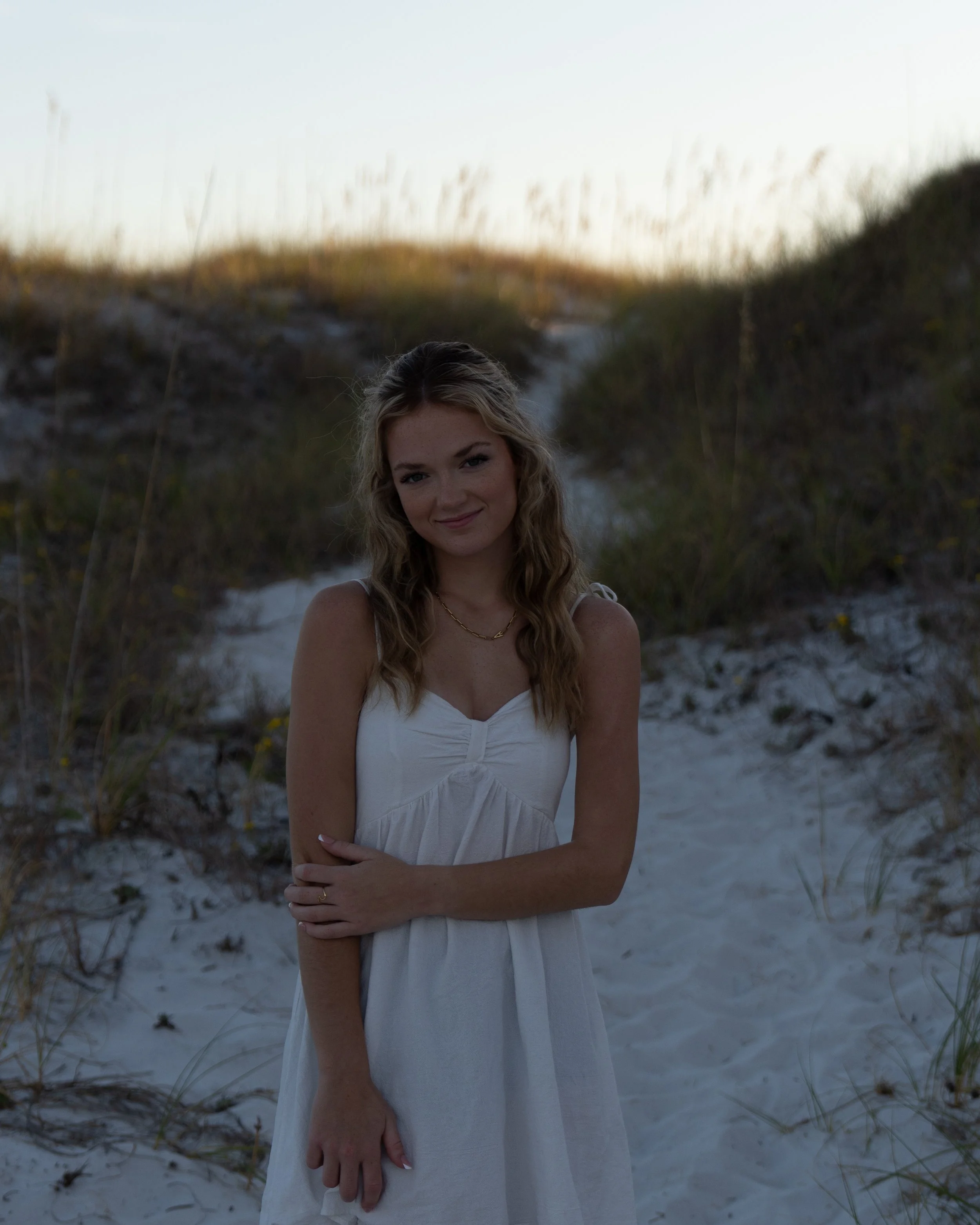 A woman with long wavy blonde hair and a white dress standing on a sandy beach with dunes and grass in the background.