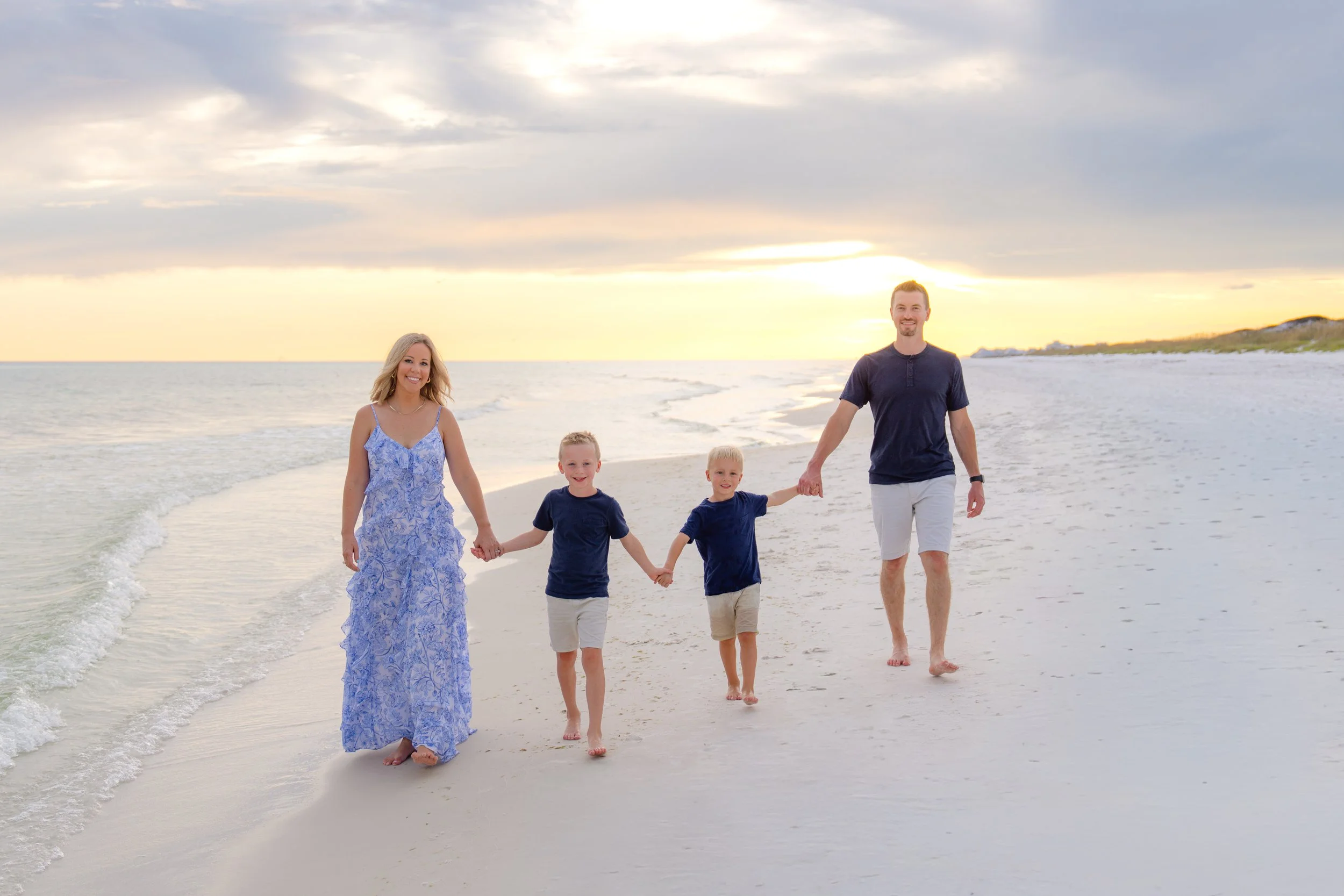 Family of four walking hand in hand along the beach at sunset, with waves and a cloudy sky in the background.