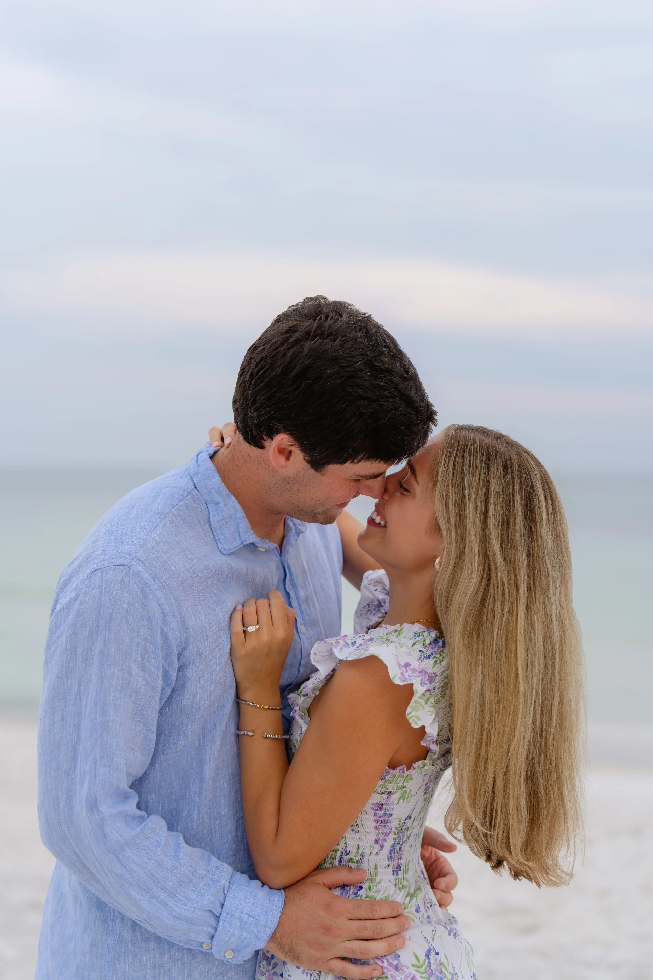 A couple embracing and smiling at each other on the beach with the ocean and sky in the background.