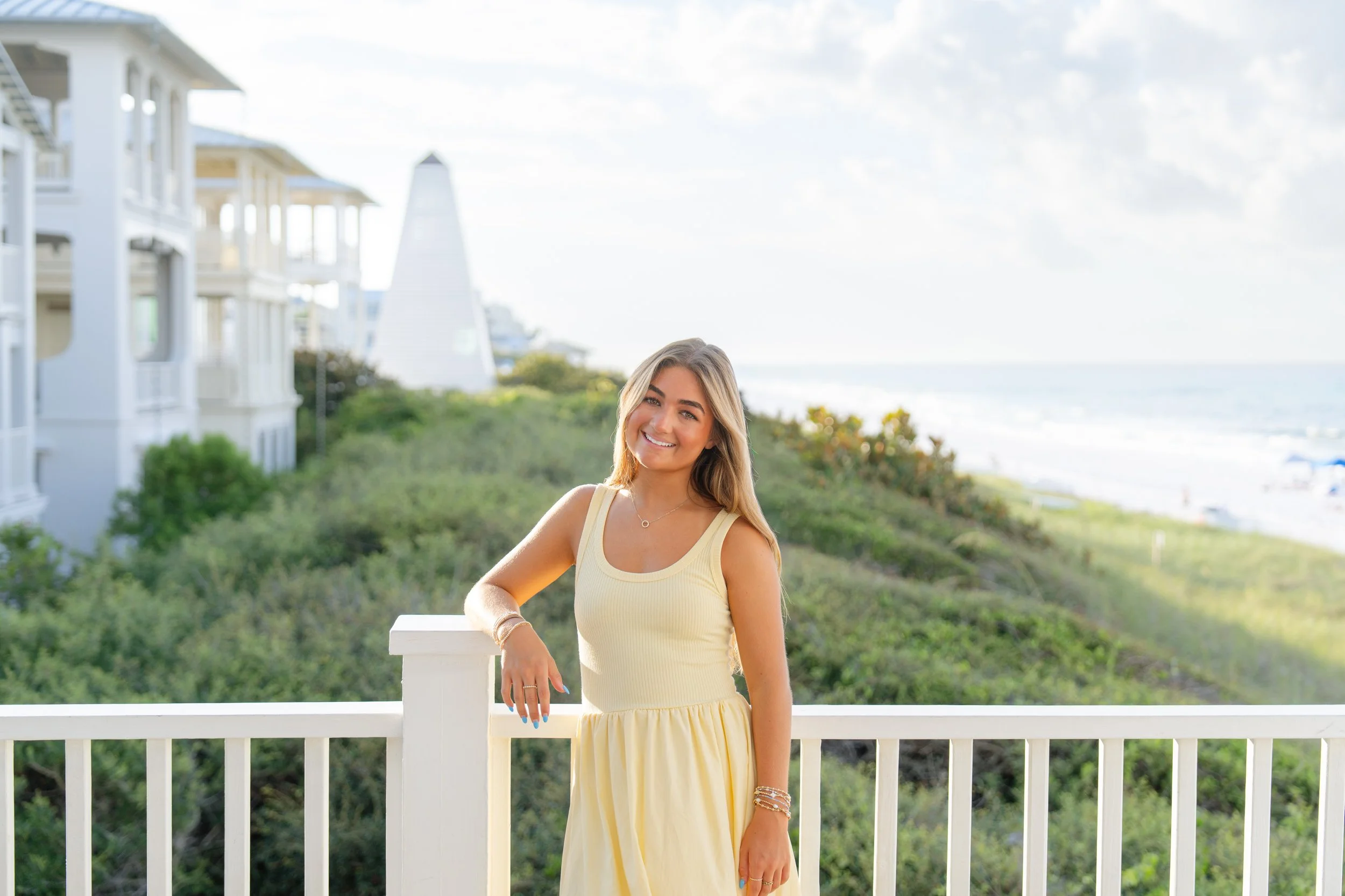 A young woman in a yellow dress smiling on a balcony with a view of greenery, some white buildings, and the beach in the background.