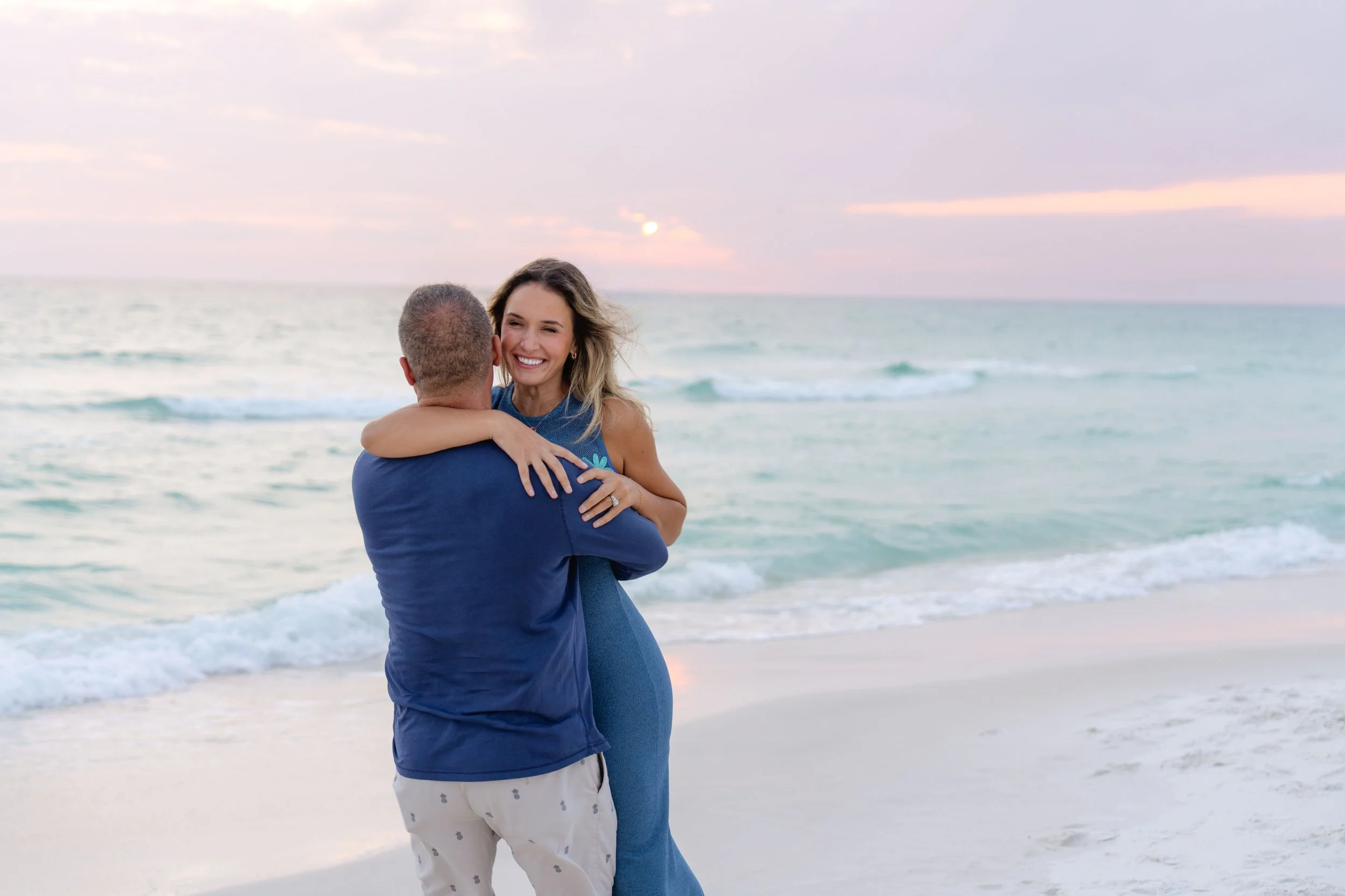 A couple hugging on a beach at sunset, with the woman smiling and looking happy.