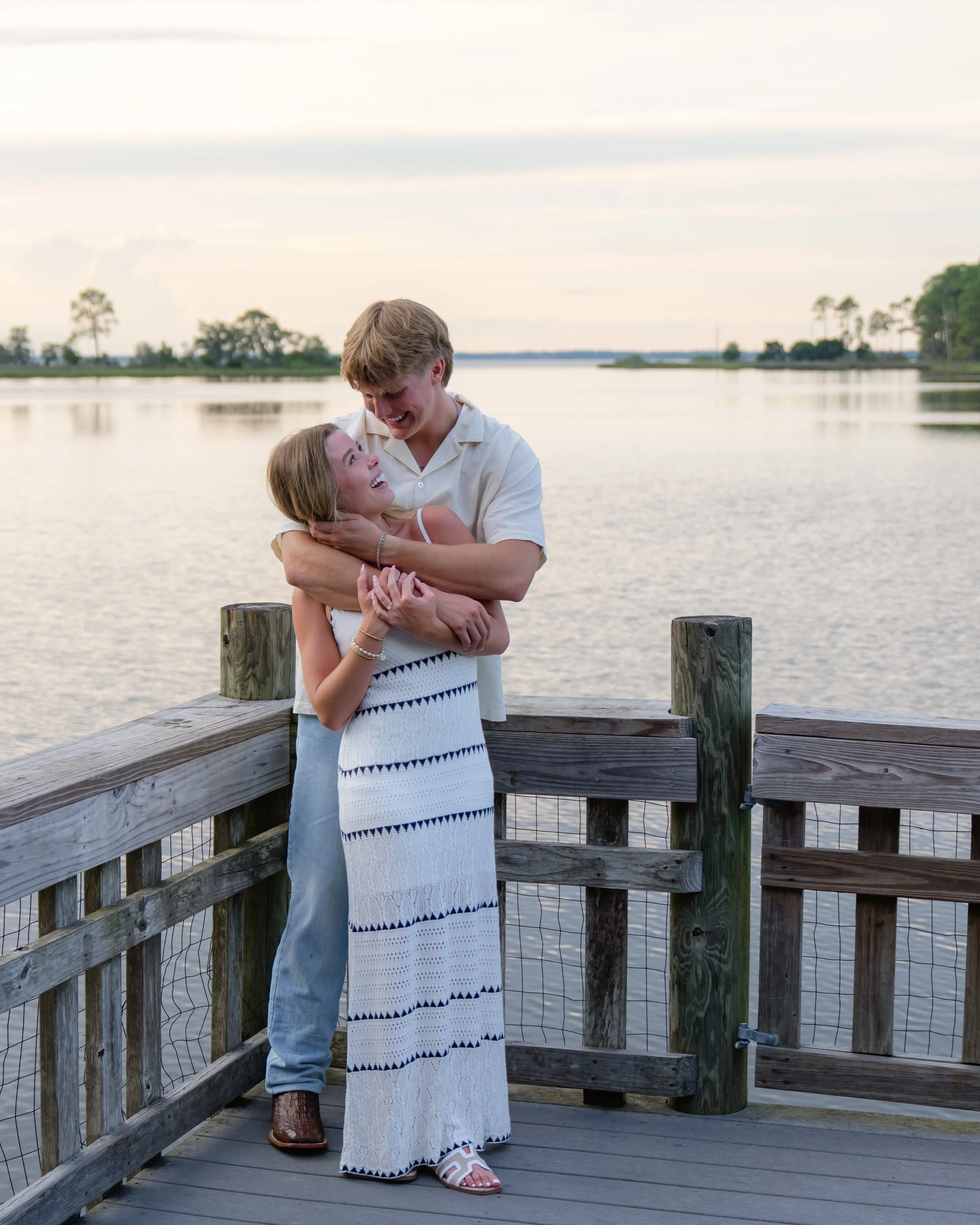 A young couple is standing on a wooden dock by a lake, embracing and smiling at each other during sunset or late afternoon.