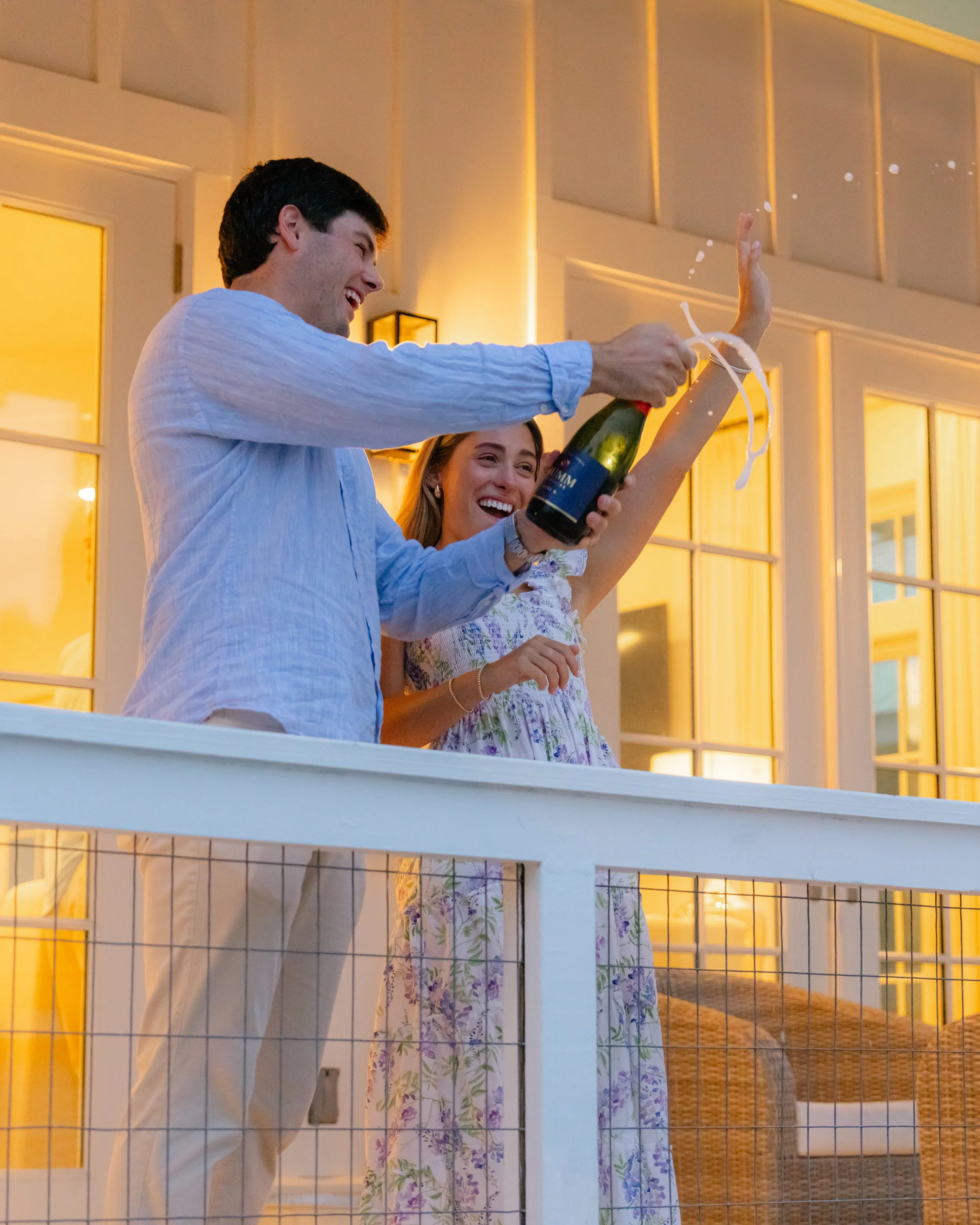 Man and woman smiling on a balcony popping a bottle of champagne and laughing. 30A Engagement Photographer