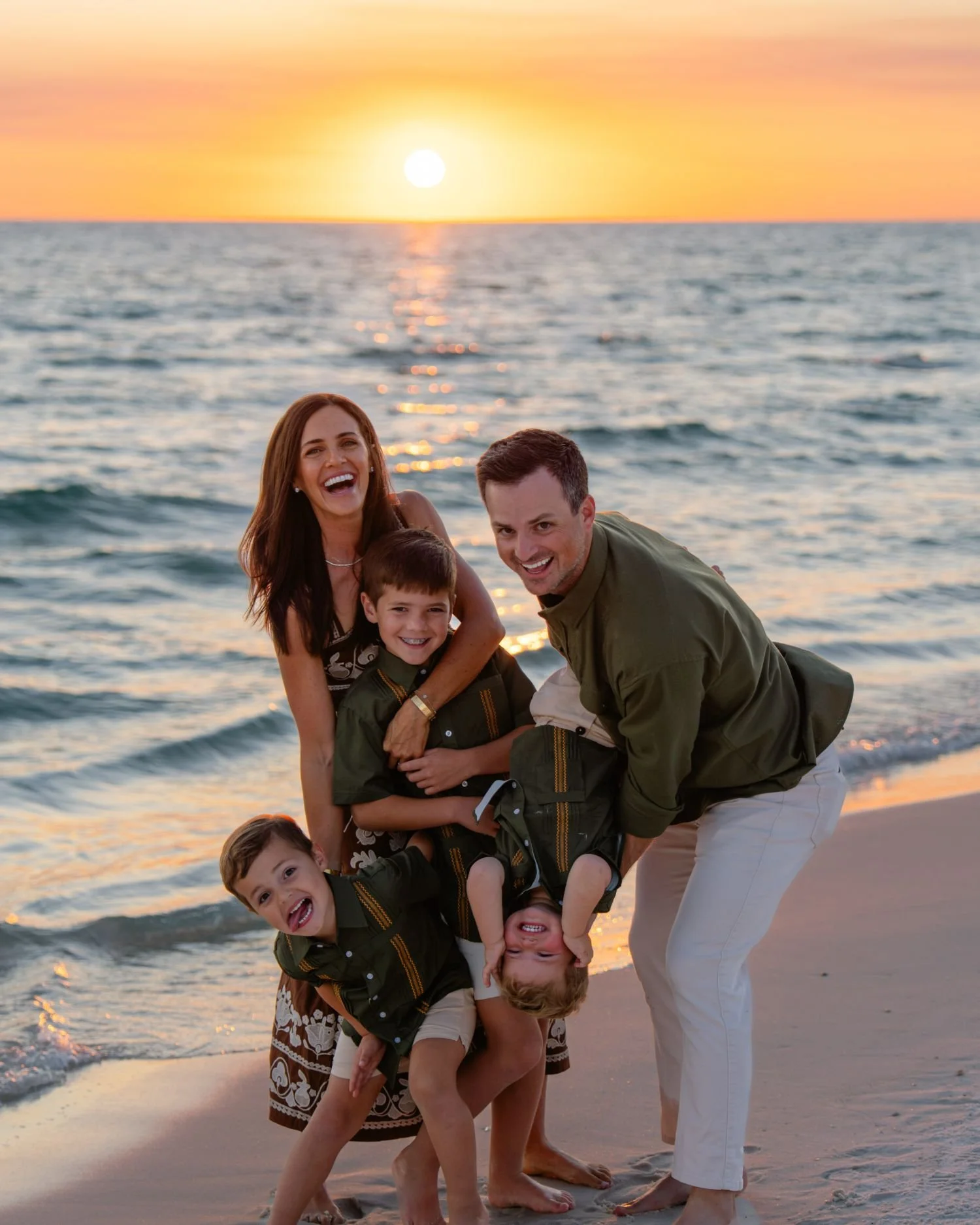A family of four enjoying time on the beach during sunset. The mother, father, and two young boys are smiling and playing near the shoreline, with the ocean and a setting sun in the background.