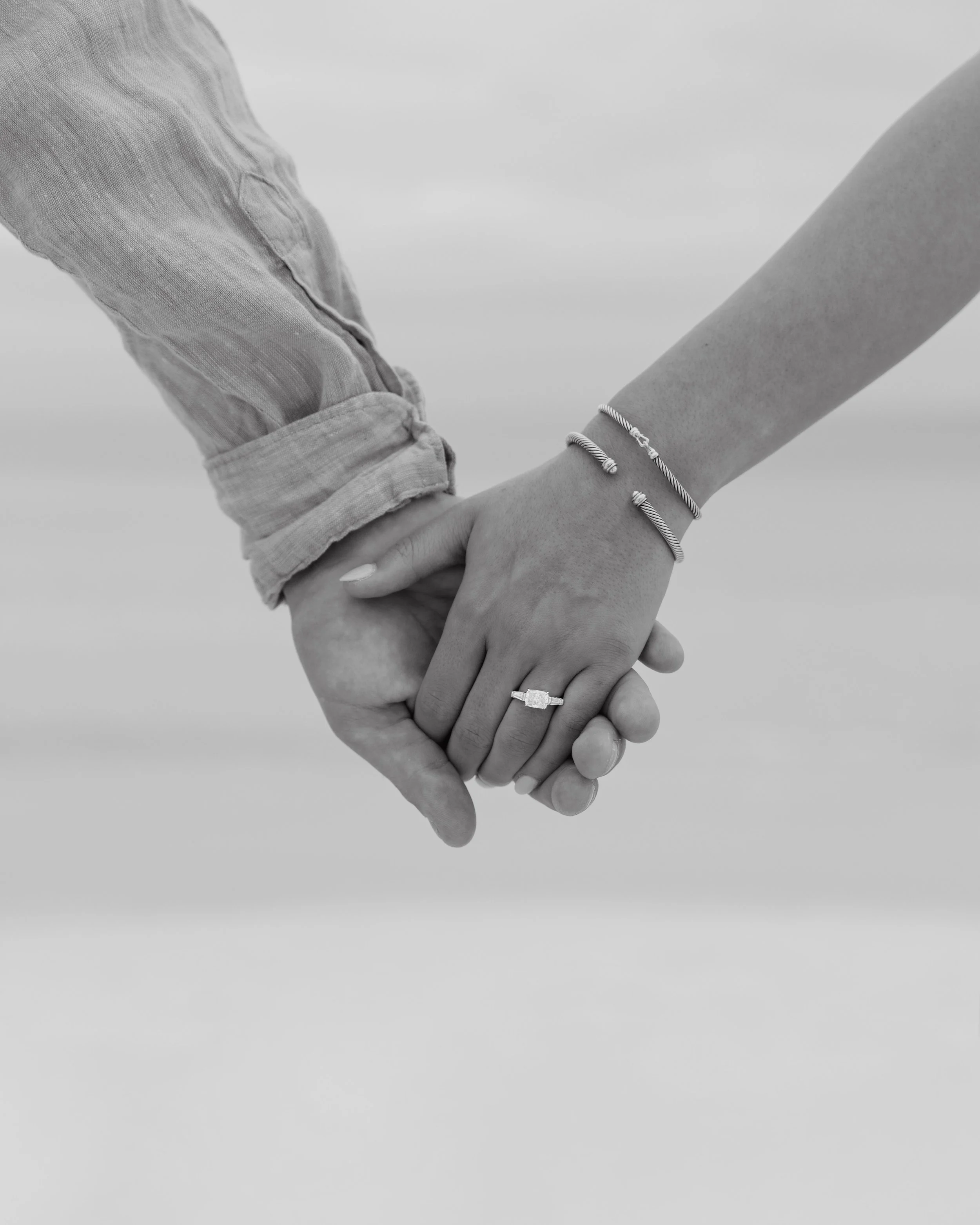 Two people holding hands, one wearing a ring and bracelets, the other wearing a rolled-up shirt, black and white photo.