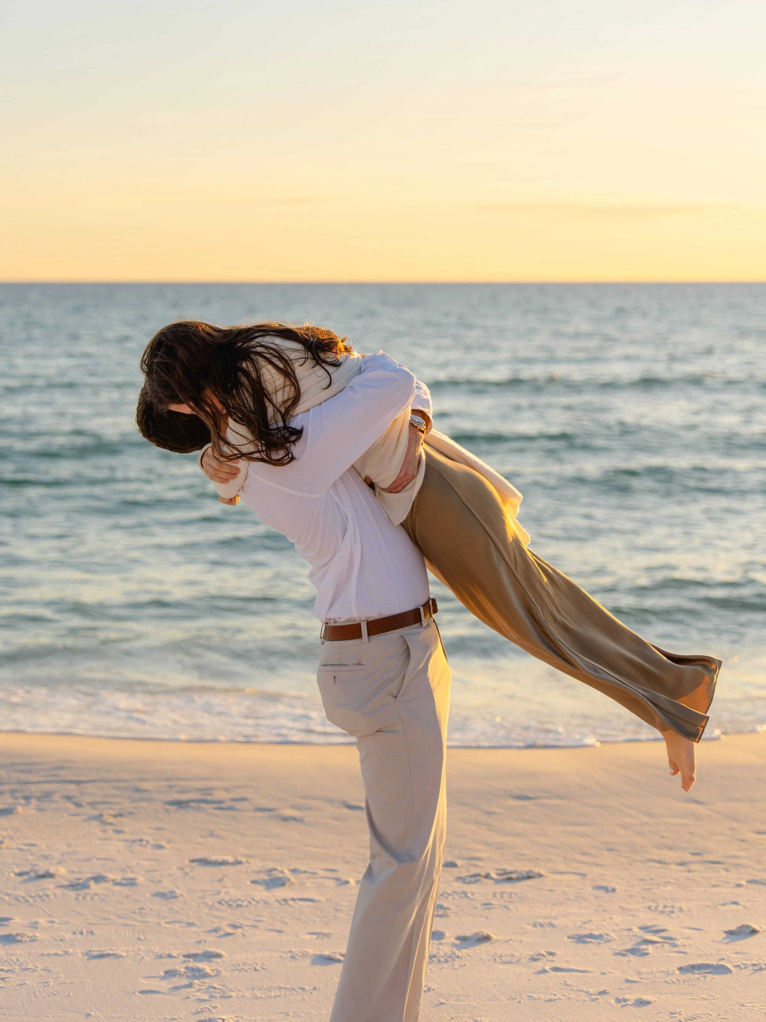 Young couple embracing on the beach at sunset with the ocean behind them. 30A Engagement Photographer