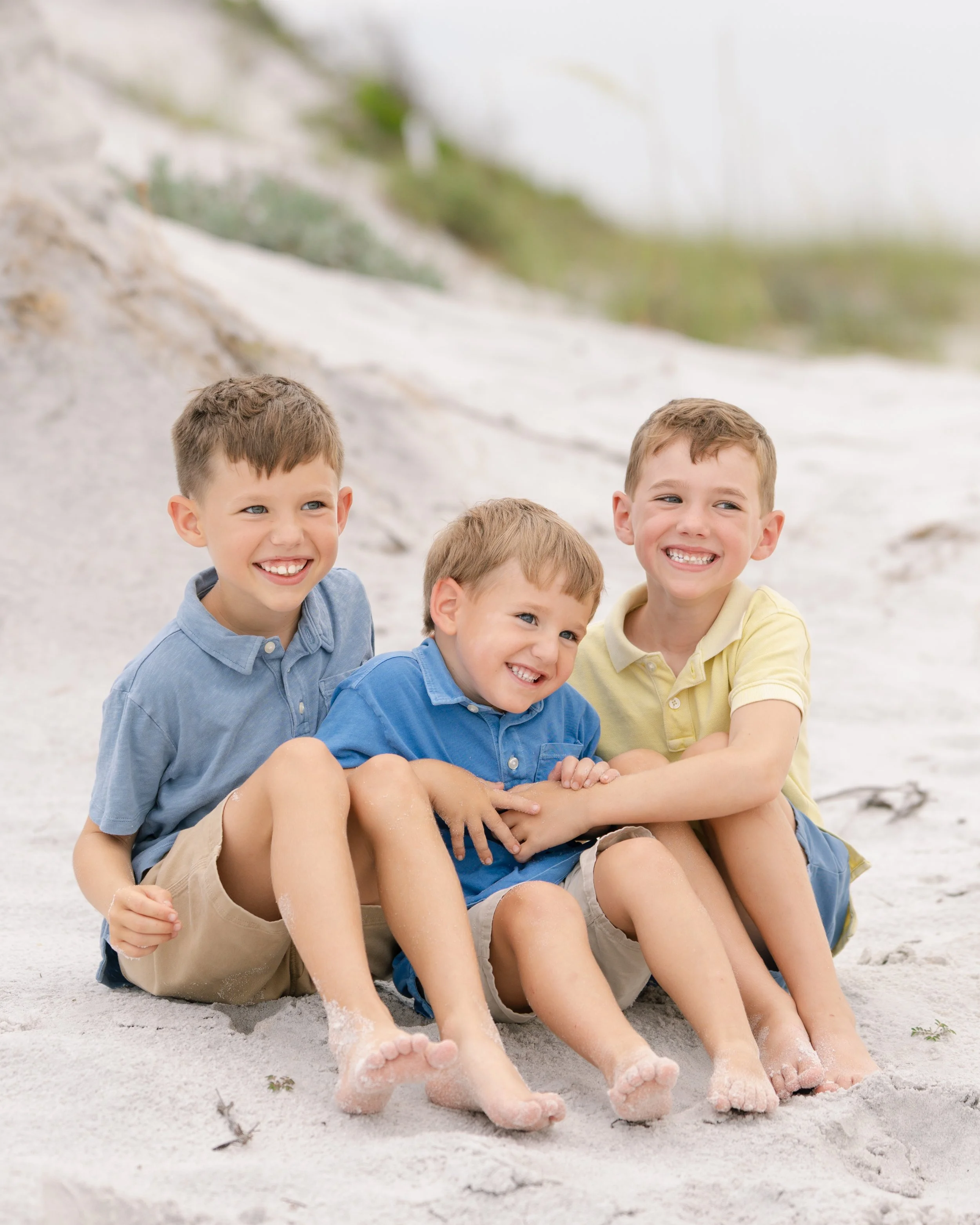 Three young boys sitting on the dunes of a beach smiling and laughing.