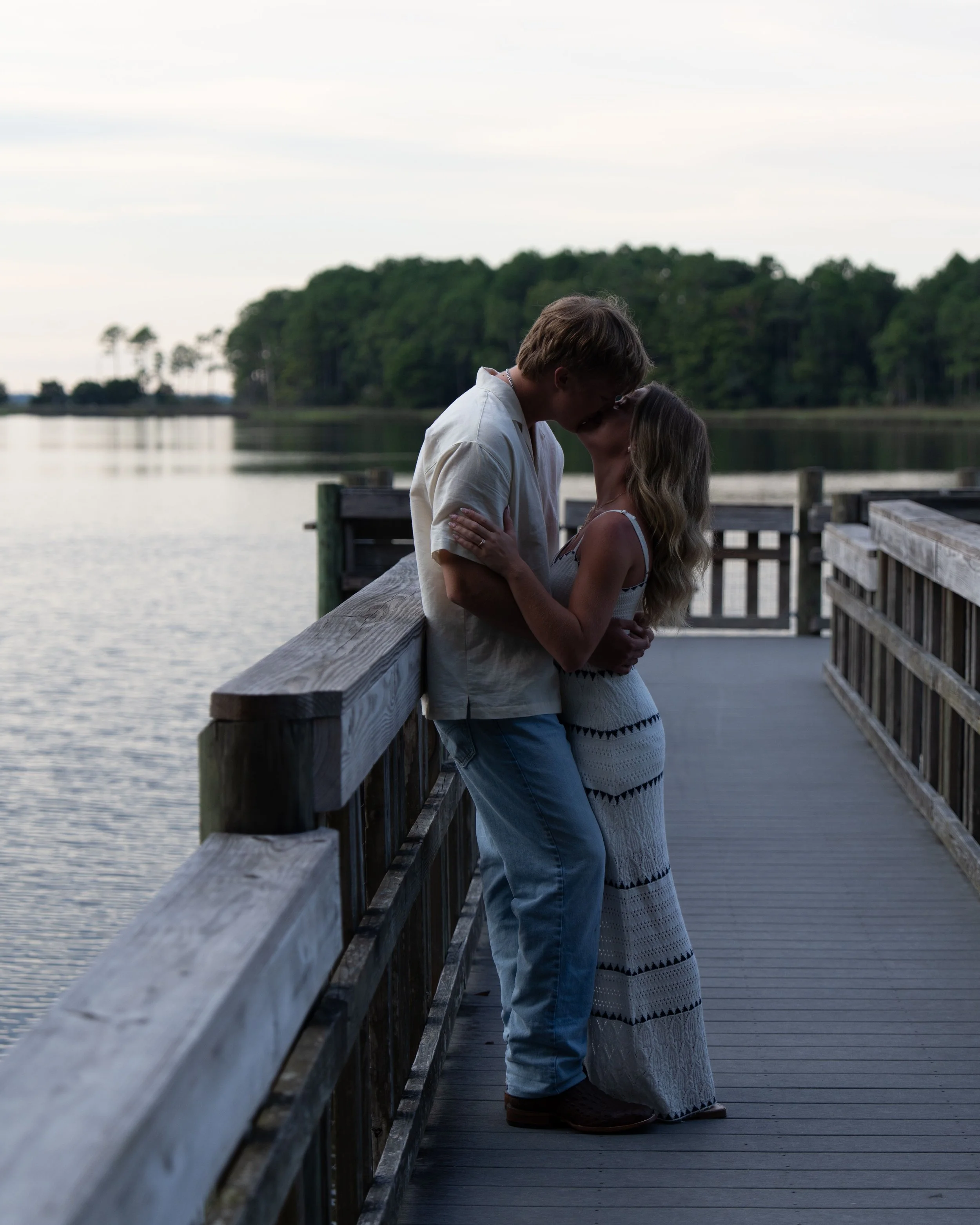 A couple is kissing on a wooden dock by a lake during sunset, with trees in the background.