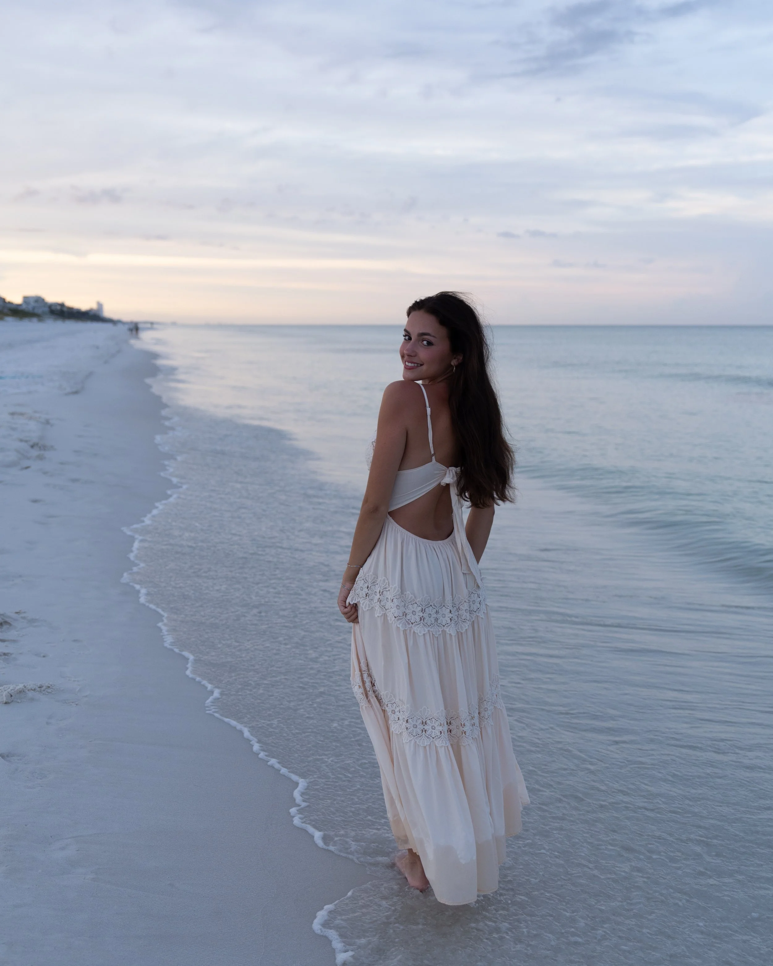 A woman in a white dress standing on the beach with her feet in the water, smiling at the camera, during sunset or dusk.