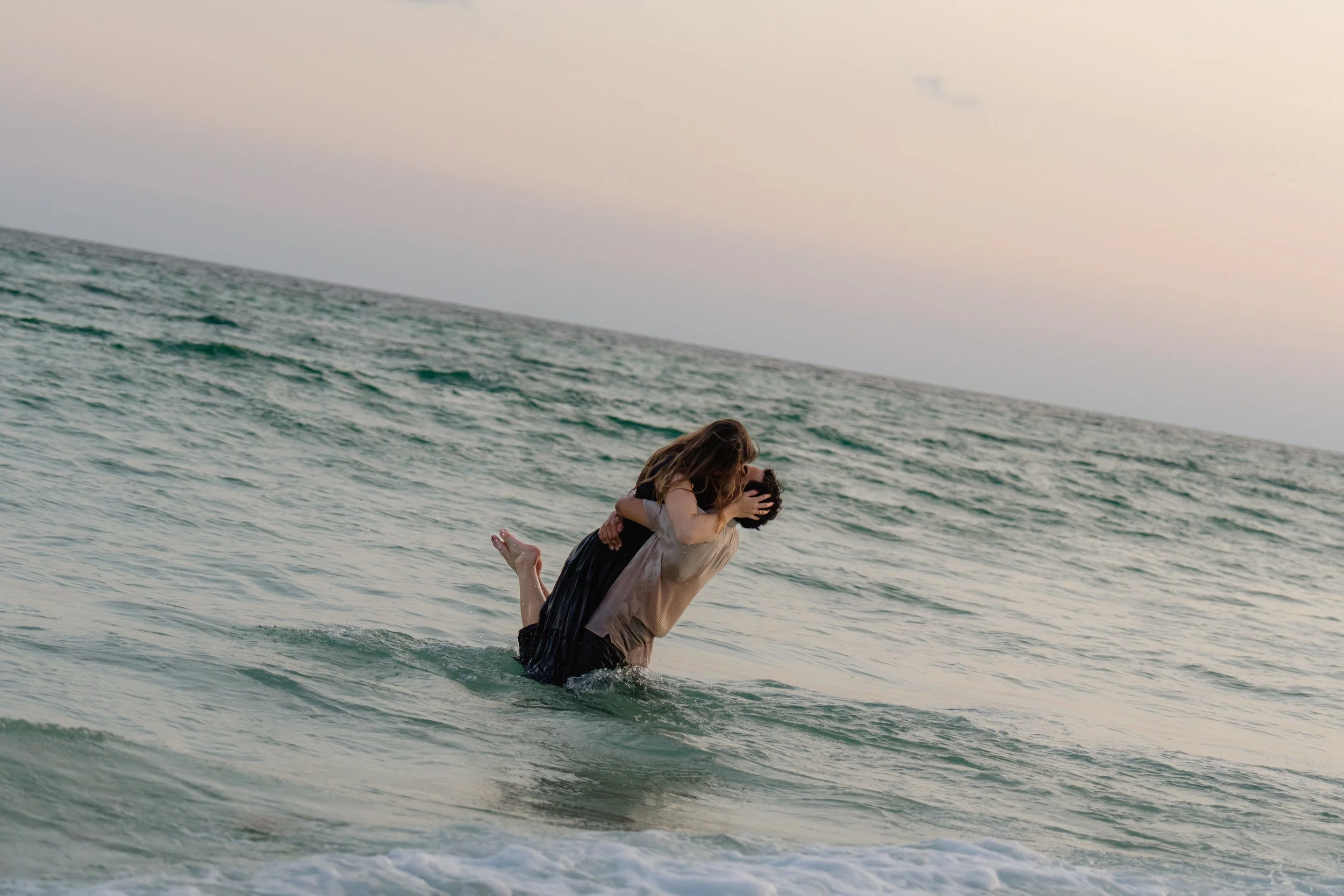 A couple playing in the ocean waves, with the man lifting the woman in the water during sunset.