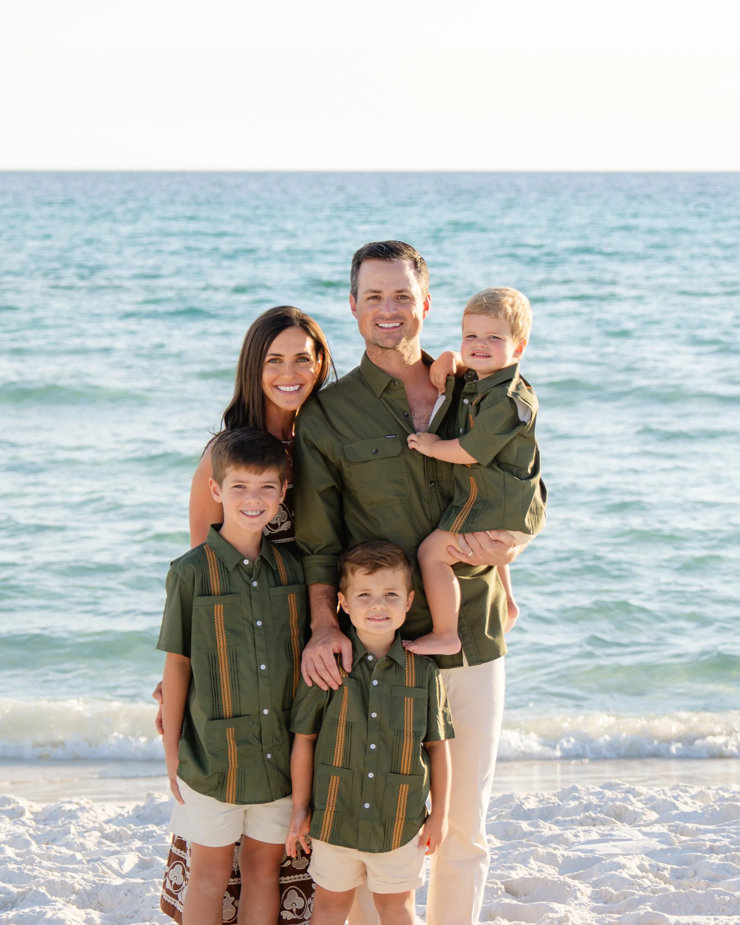 A family of six enjoying a day at the beach, standing on white sand with the ocean in the background. The family includes two adults and four children, all dressed in matching green shirts and light-colored shorts.