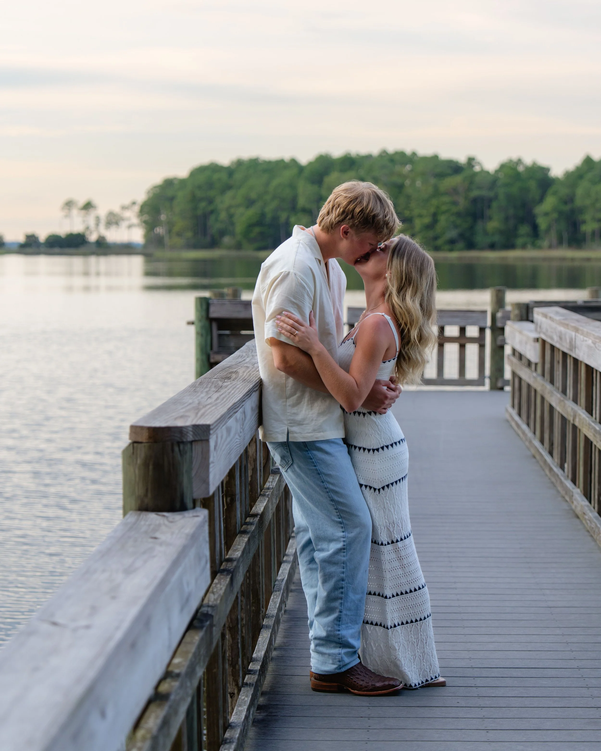 A romantic couple sharing a kiss on a wooden pier by a lake during sunset, with a dense green forest in the background.